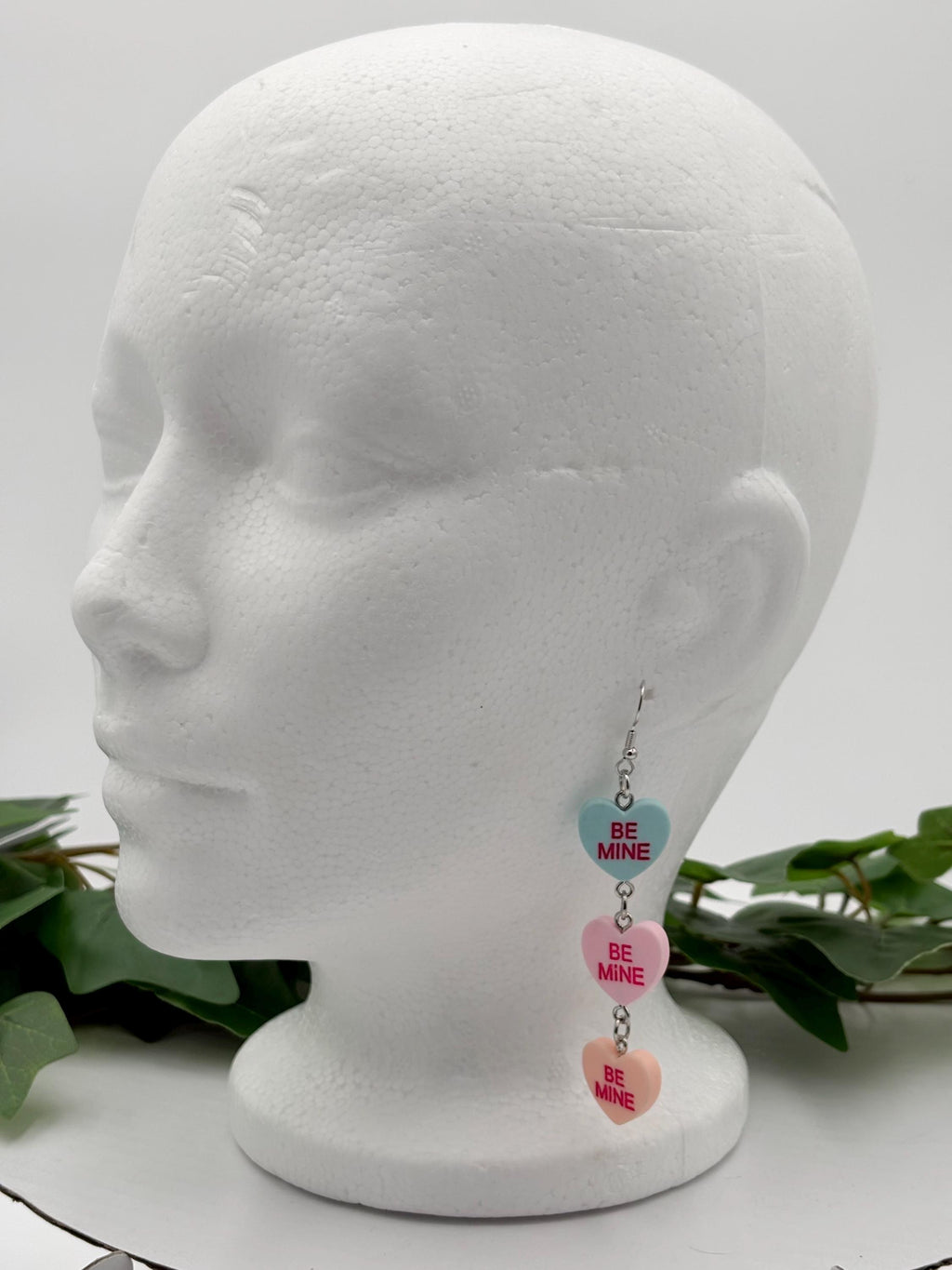 Heart-shaped dangle earrings on a mannequin head with green leaves in the background