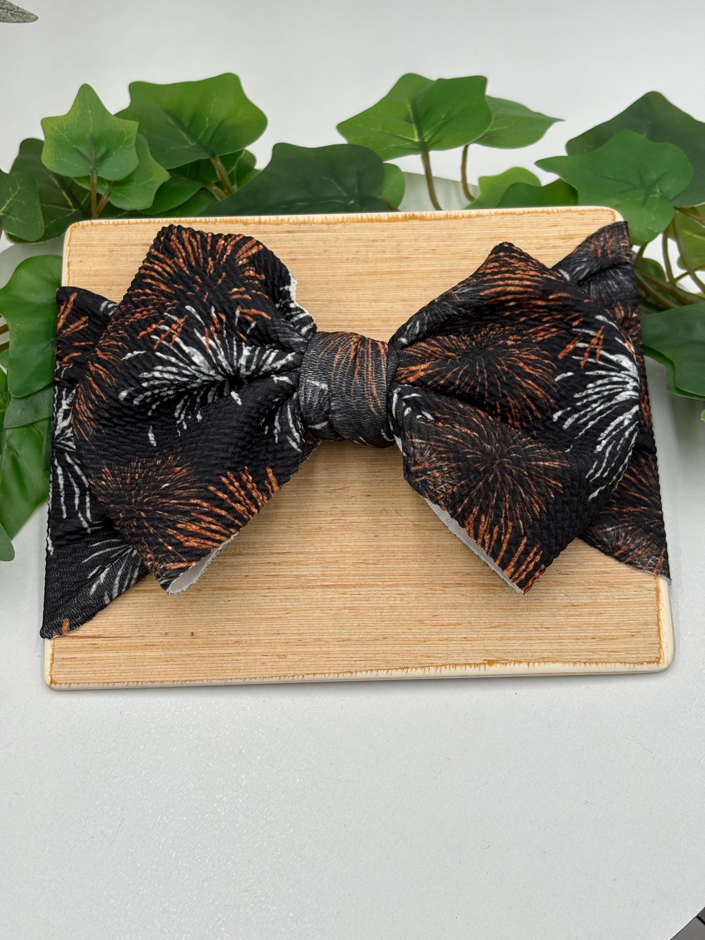 Black Headband with a decorative orange and white fireworks themed big bow on a wooden board against a white background