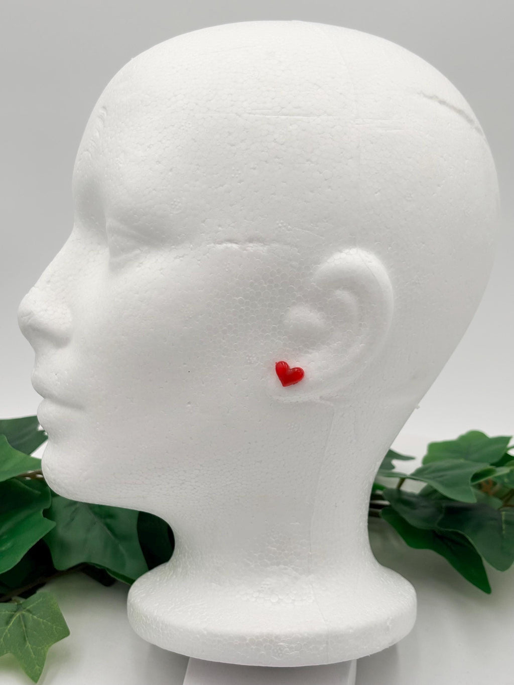 Red heart-shaped earrings on a white mannequin head with green leaves in the background