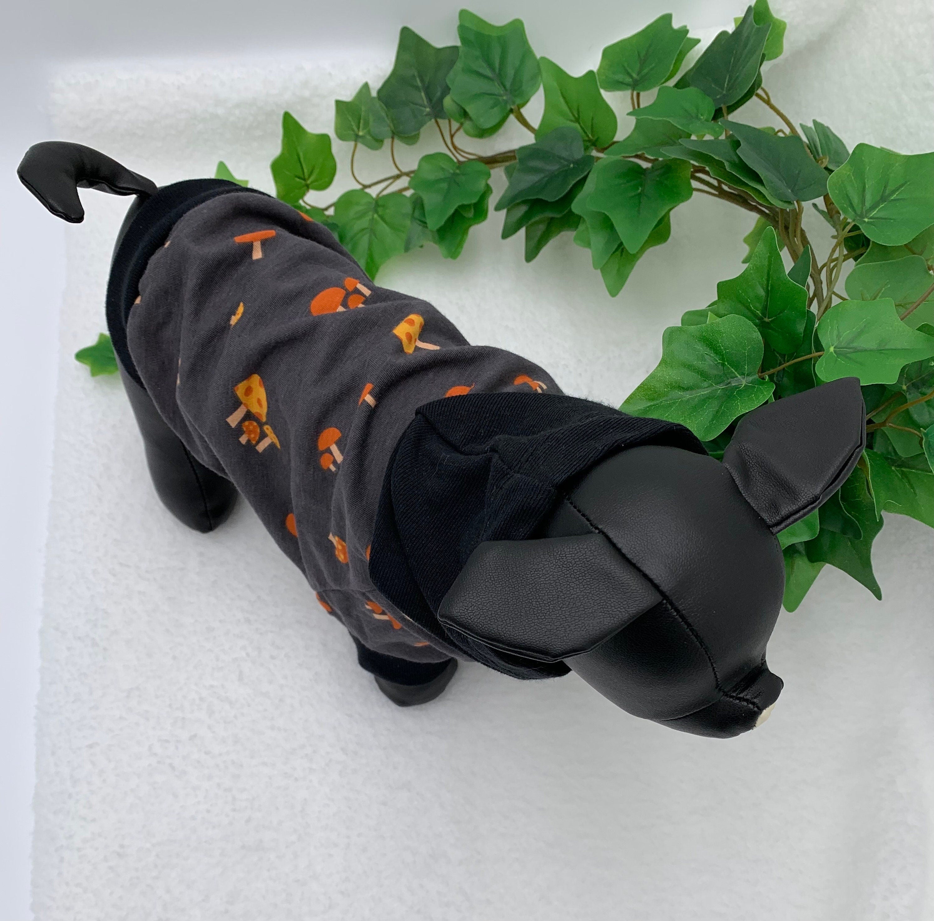 Birdseye view of Black dog mannequin wearing a gray mushroom patterned hoodie with hoodie down, standing next to a potted plant on a white surface.