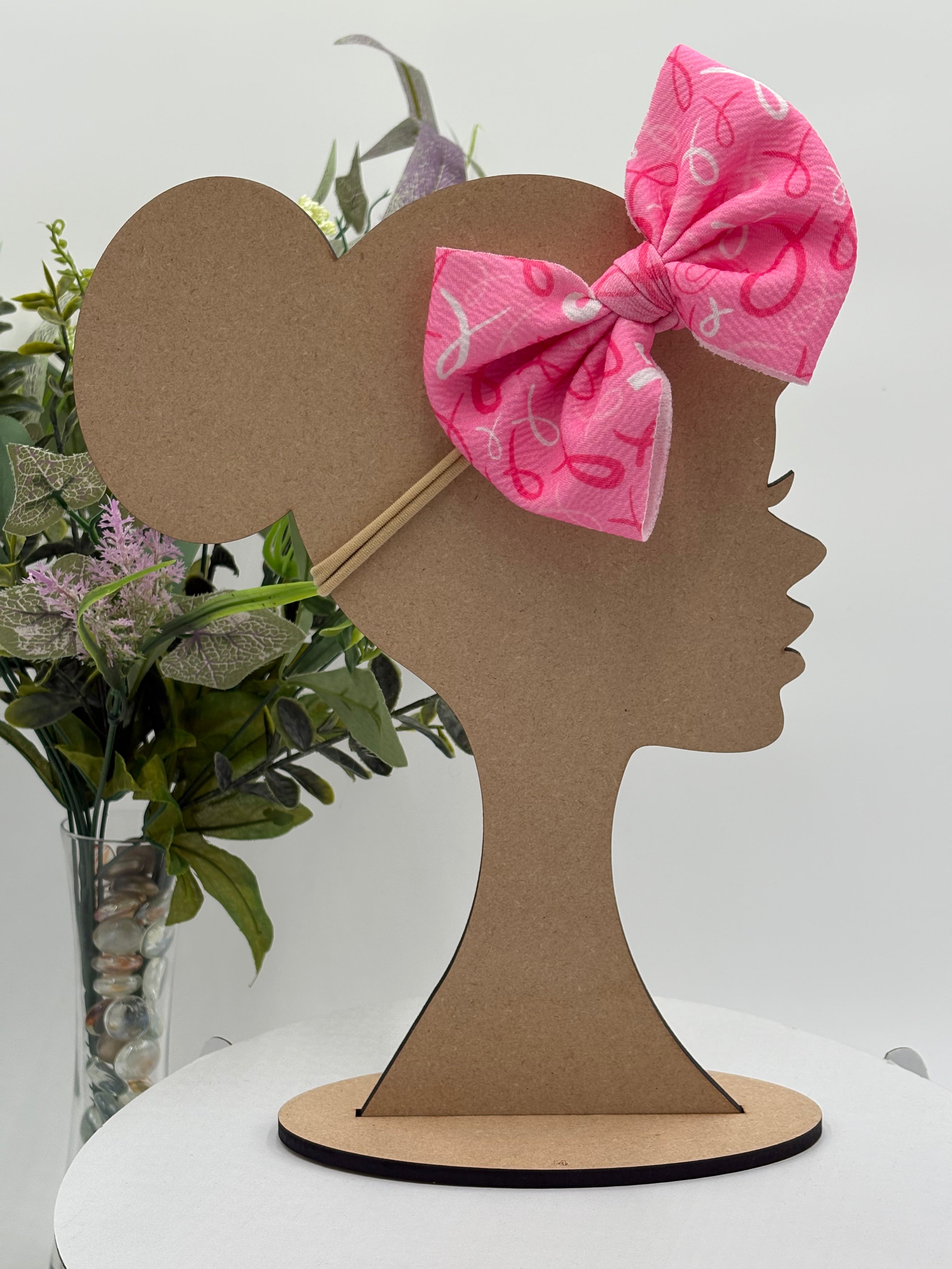 Pink Breast Cancer awareness themed big bow nylon headband on a mannequin head with a white background and greenery 