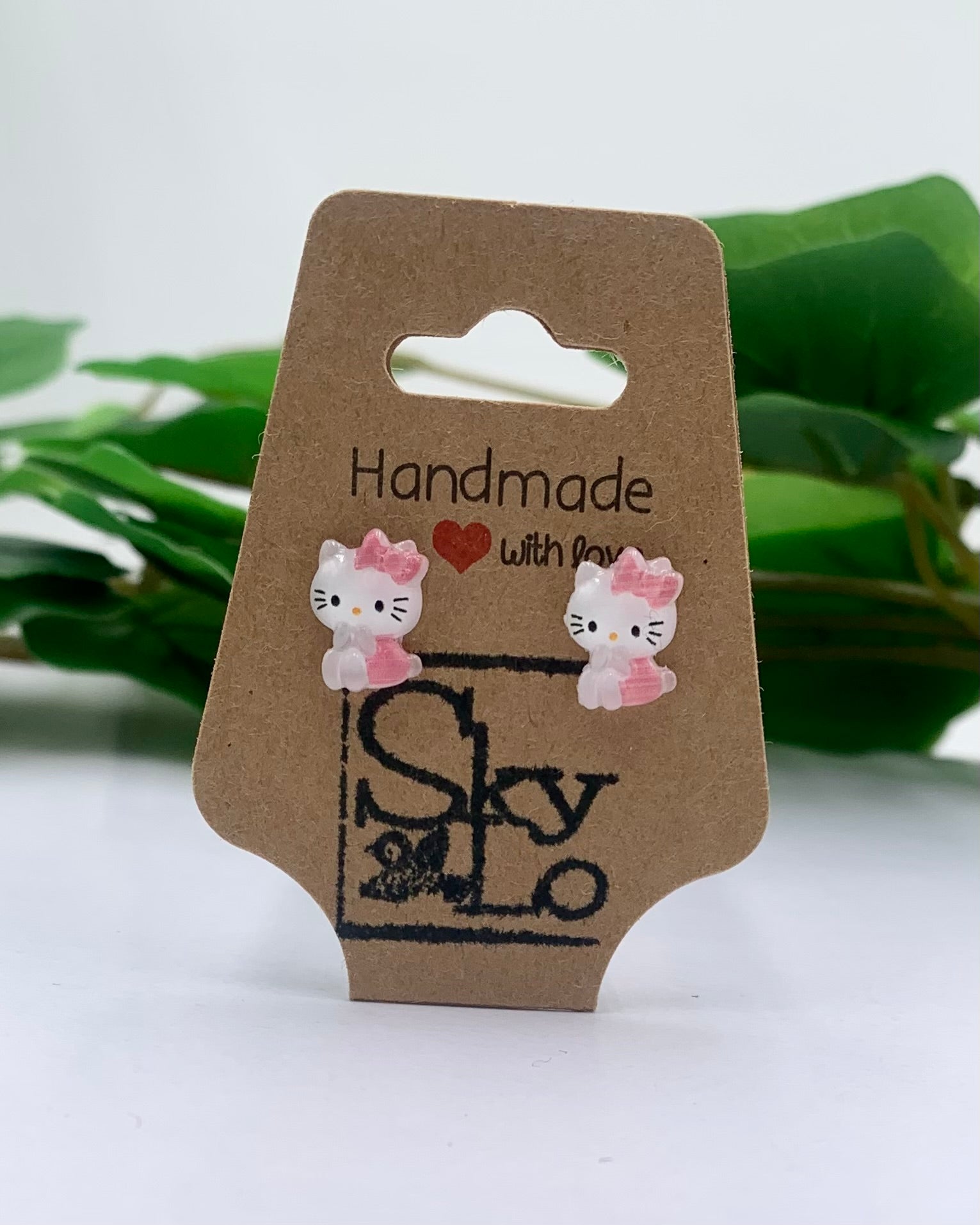 Cartoon cat earrings on a 'Handmade with Love' branded card with green leaves in the background.
