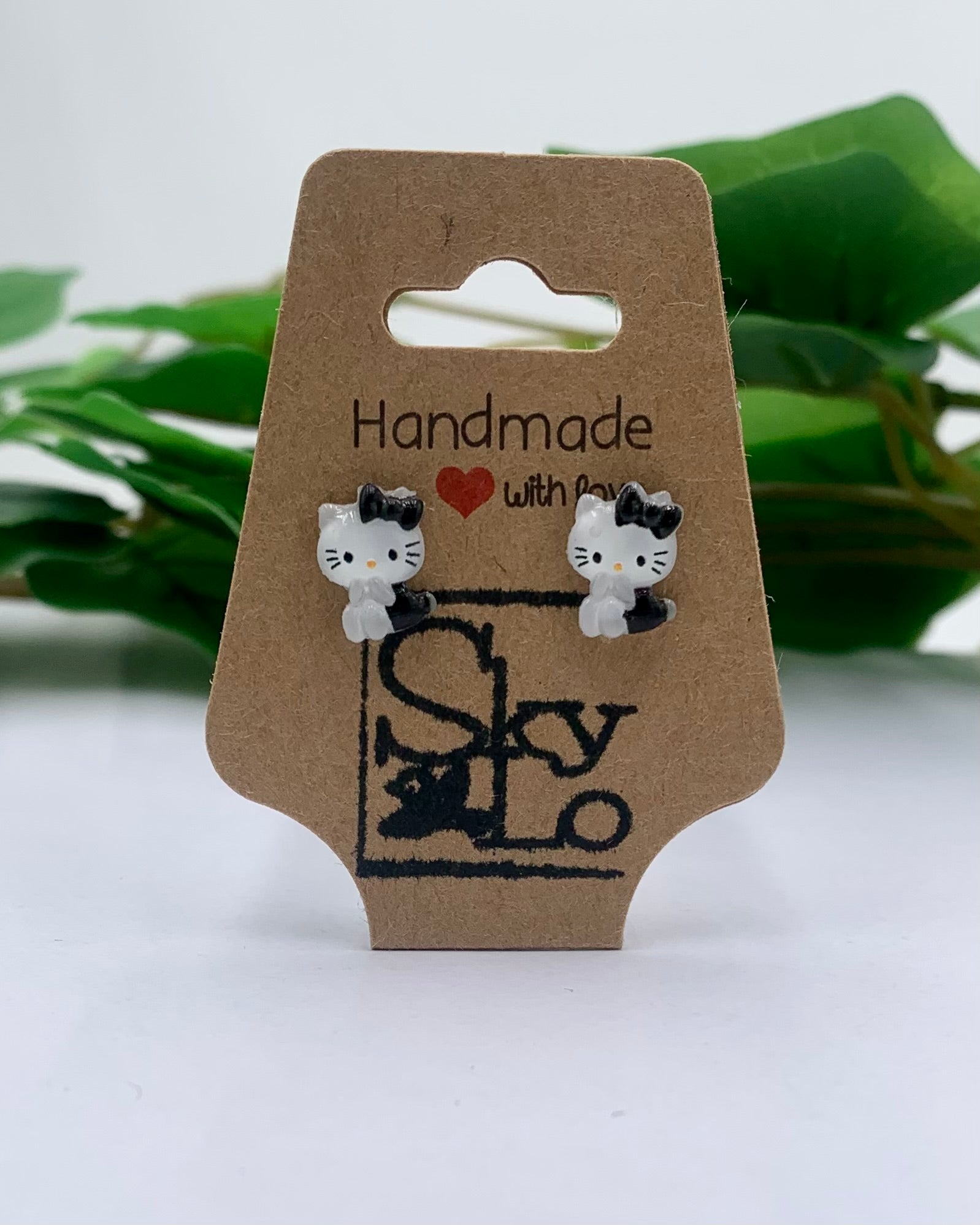 Pair of cat-shaped earrings on a 'Handmade with Love' branded card with green leaves in the background.