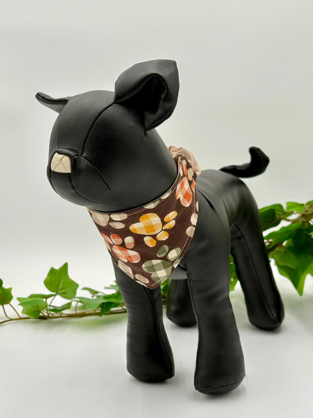 Black dog mannequin wearing a brown with plaid paw prints scrunchie bandana on a white background with green leaves.