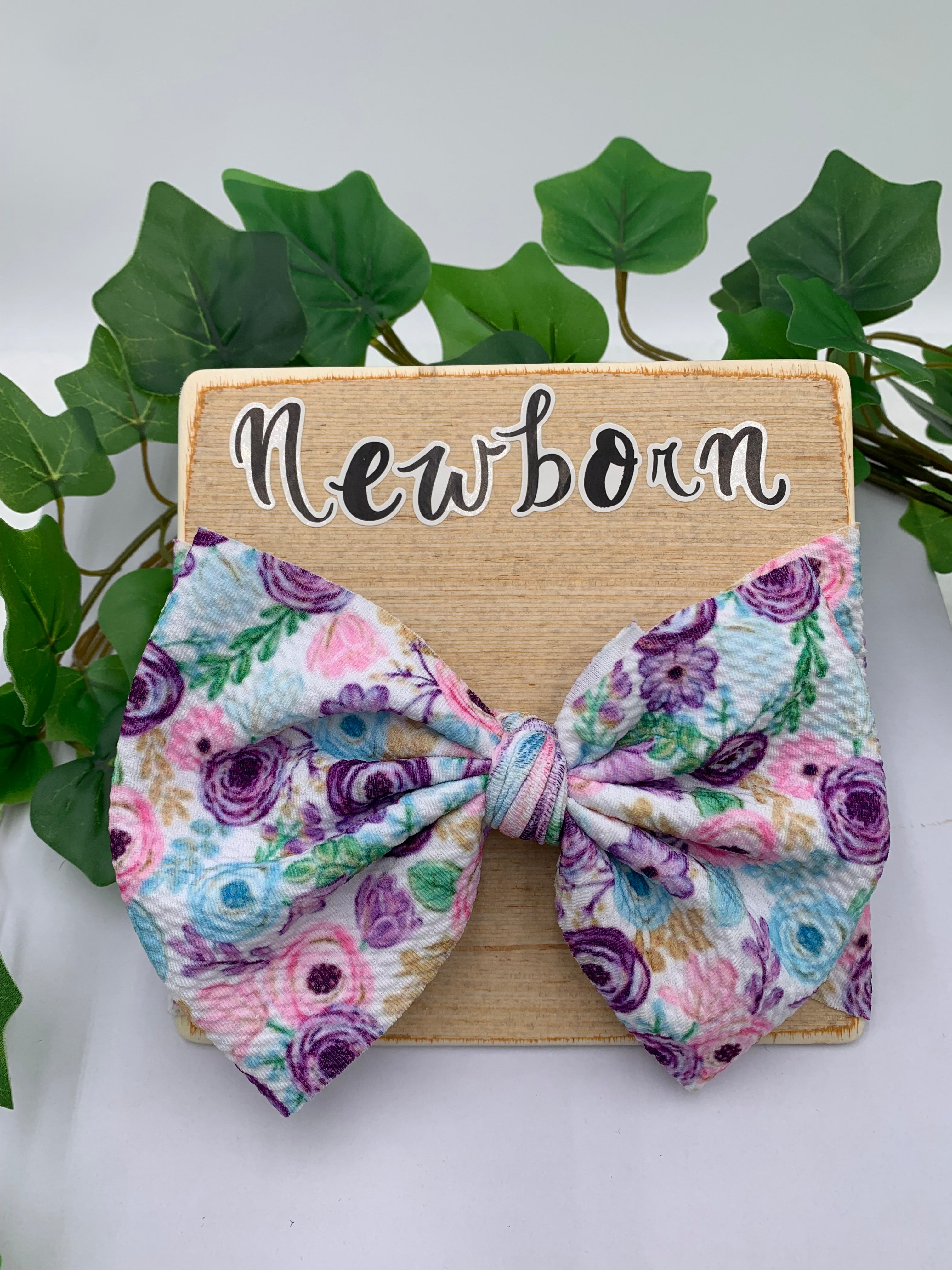 Floral big bow headband on a wooden board with 'Newborn' text, surrounded by green leaves.