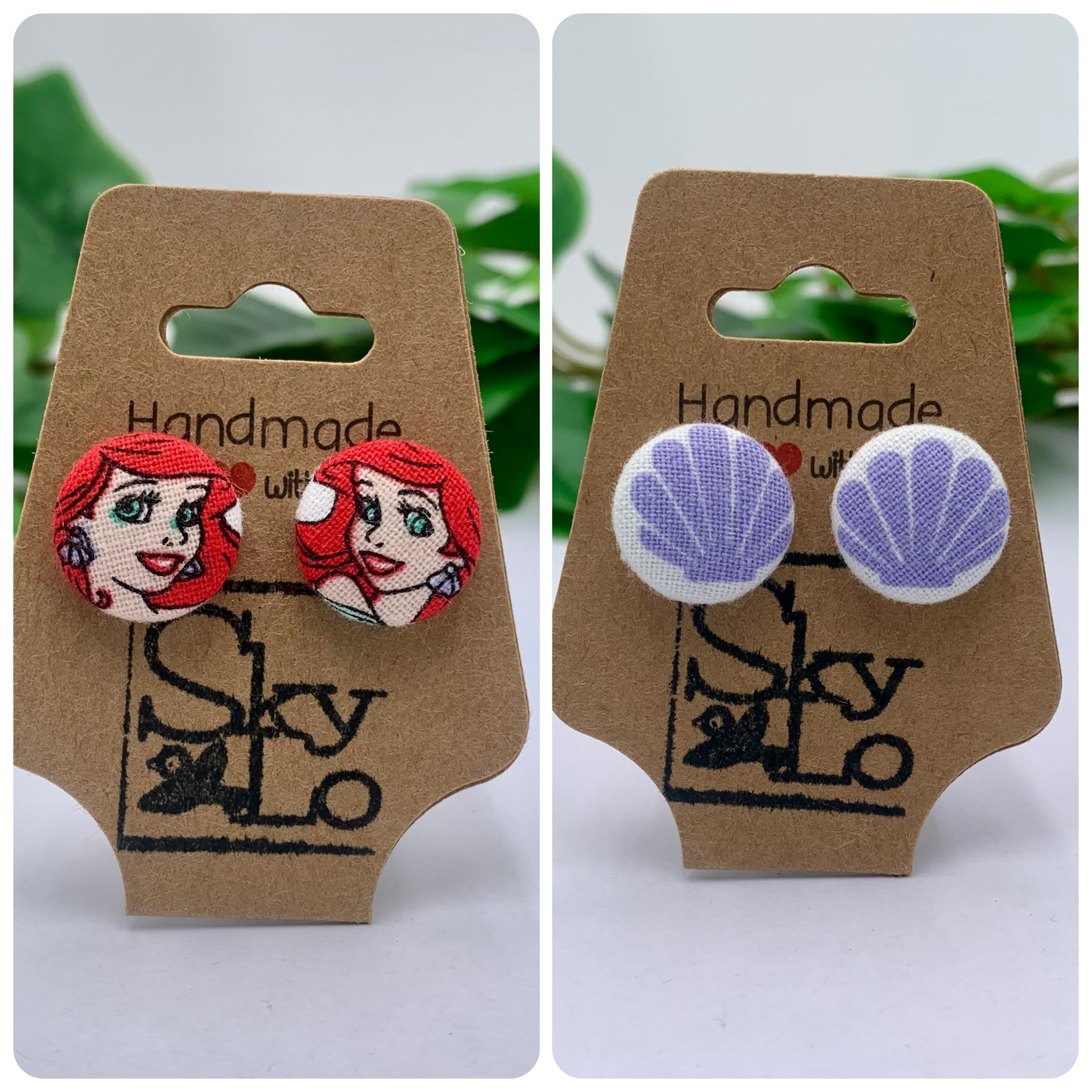 Two pairs of earrings, Ariel and seashells, on branded cardboard cards with a blurred green leafy background.