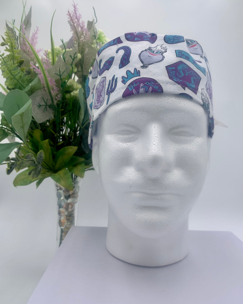 Colorful Little Mermaid Ursula scrub cap on a mannequin head with flowers, against a white background