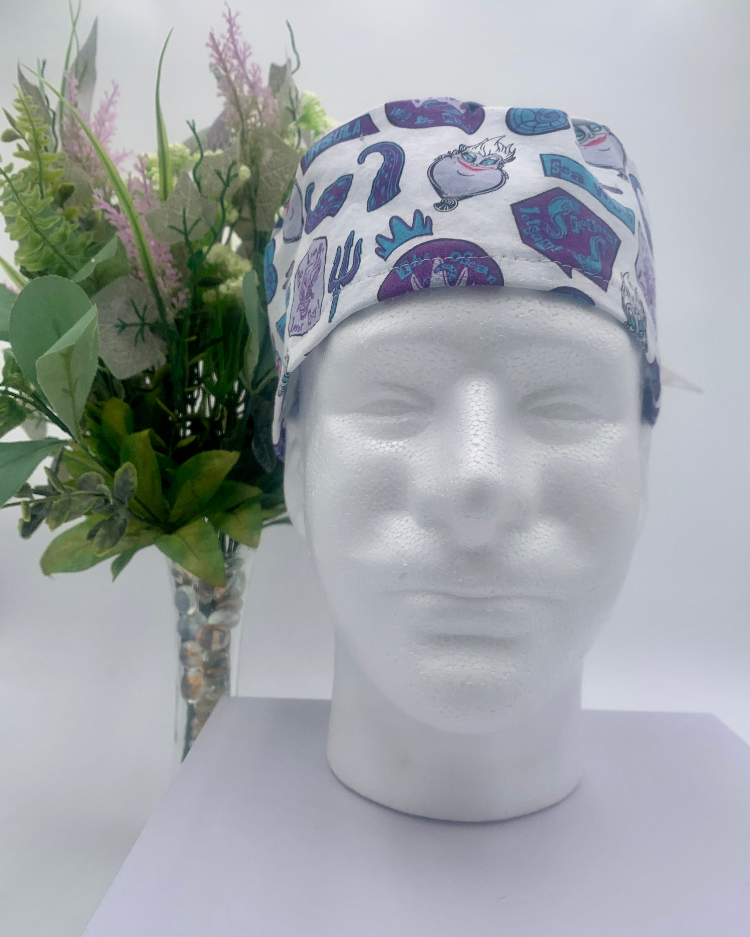 Colorful Little Mermaid Ursula scrub cap on a mannequin head with flowers, against a white background