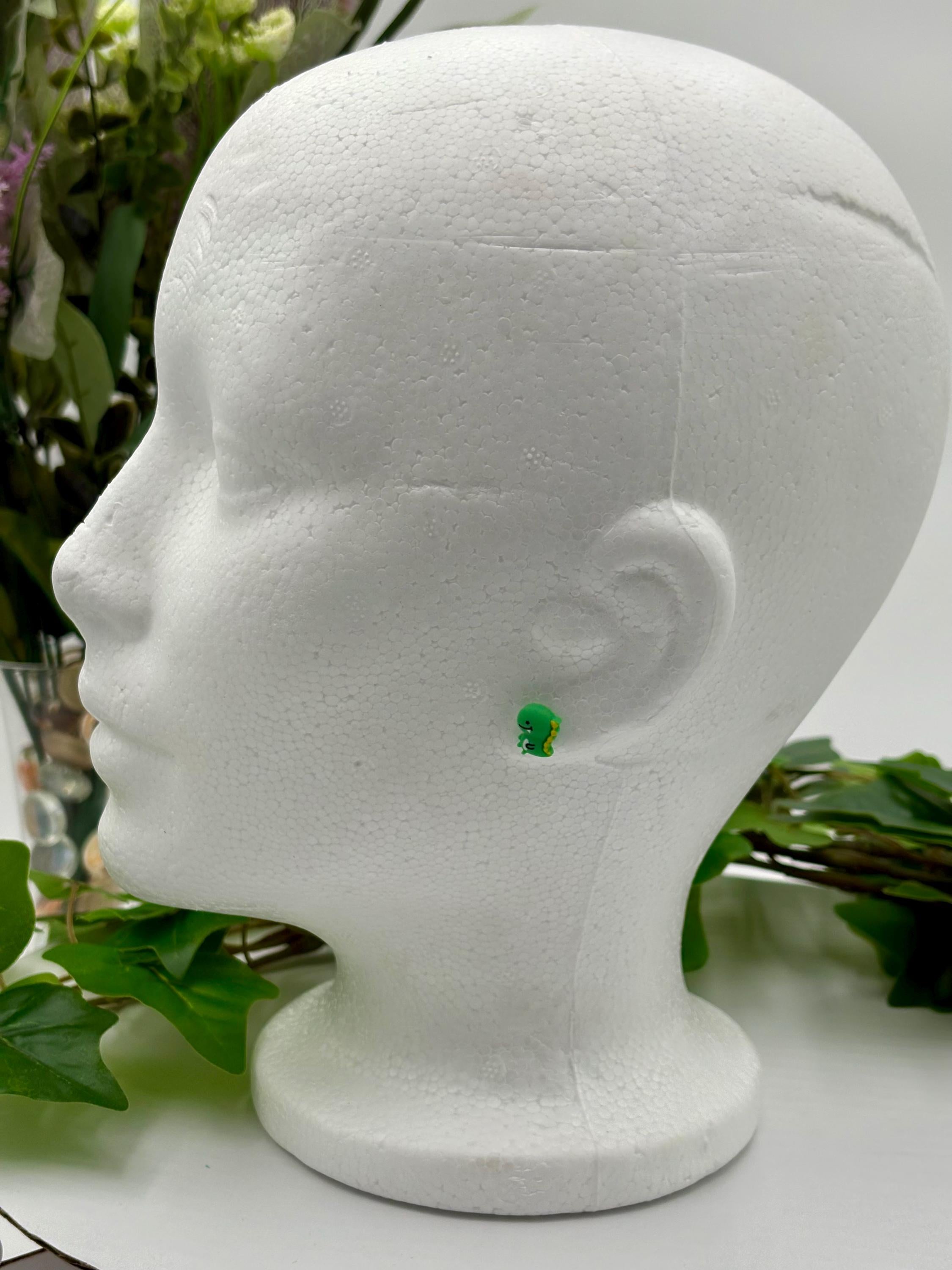 Green dinosaur shaped earring on a white mannequin head with leaves in the background