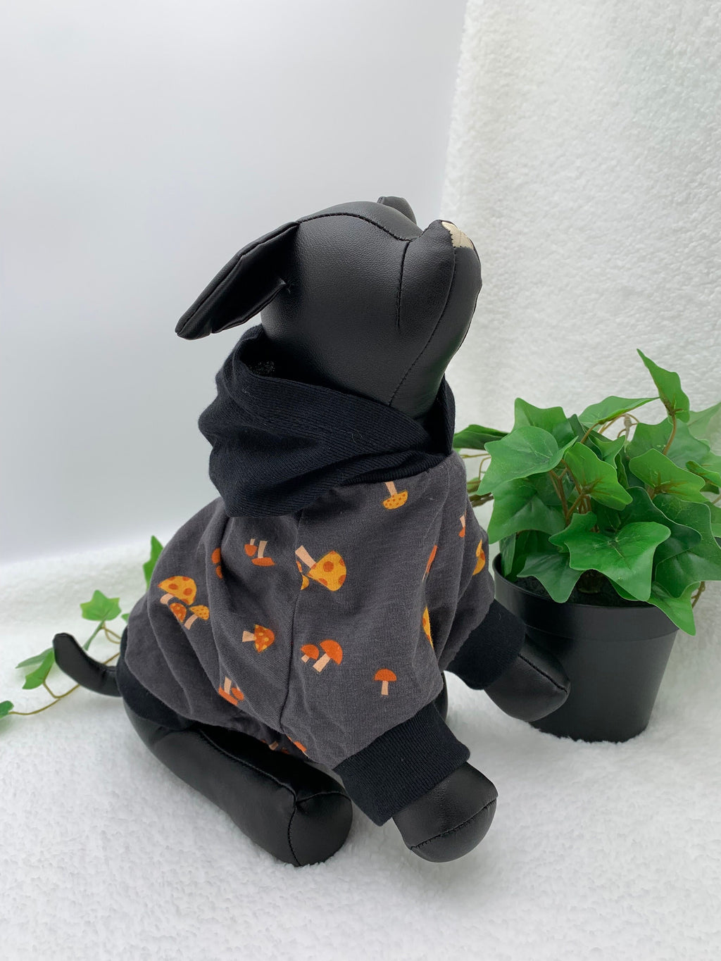 Black dog mannequin wearing a gray mushroom patterned hoodie with hoodie down, sitting next to a potted plant on a white surface.