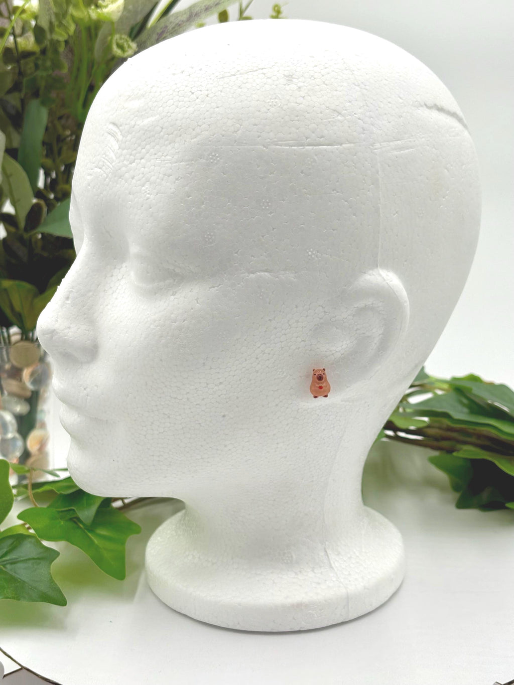 White mannequin head with a small capybara with red fruit earring on a white surface with green leaves in the background.