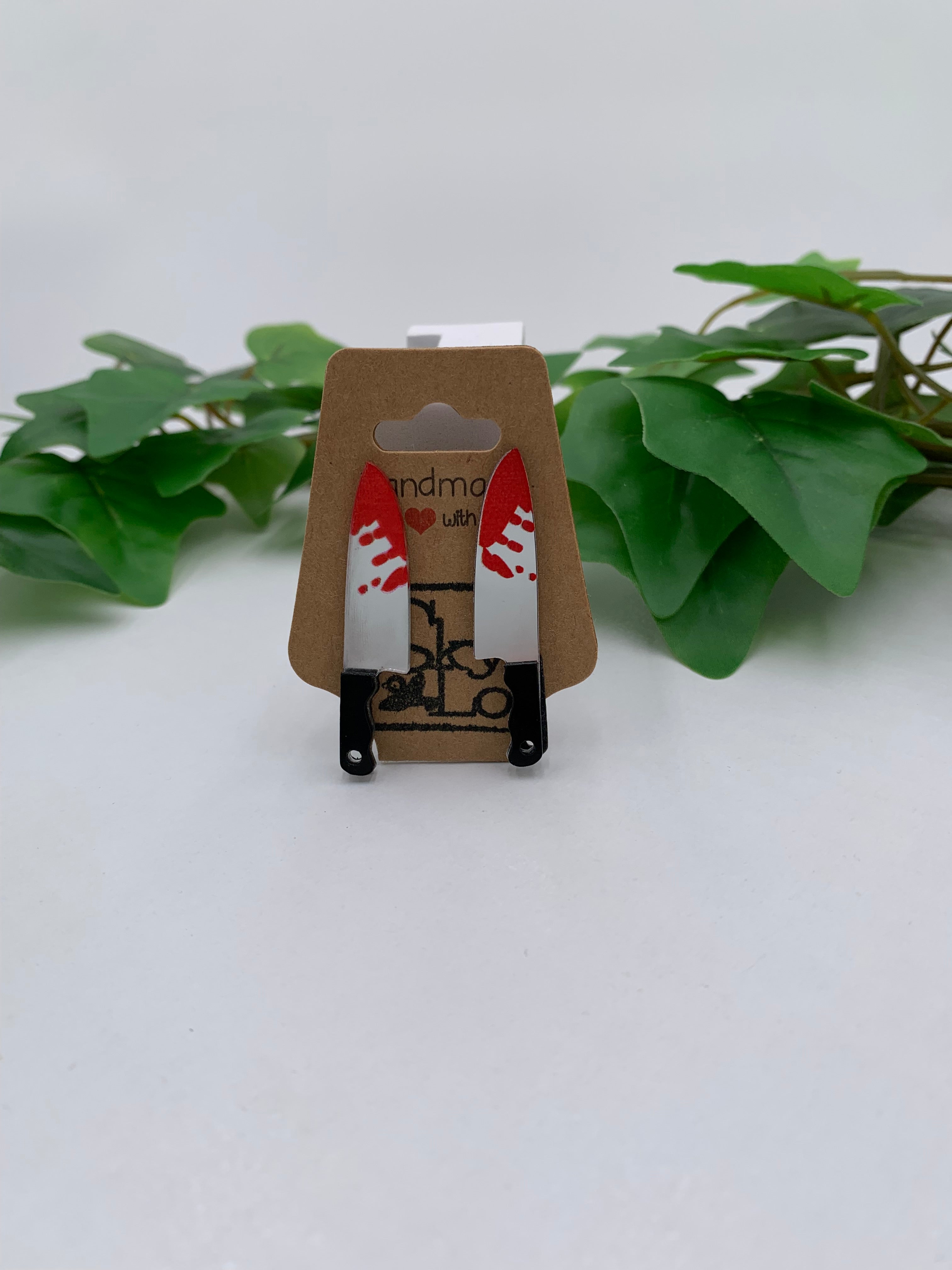 Small bloody butcher knife earrings on a kraft branded card with green leaves in the background
