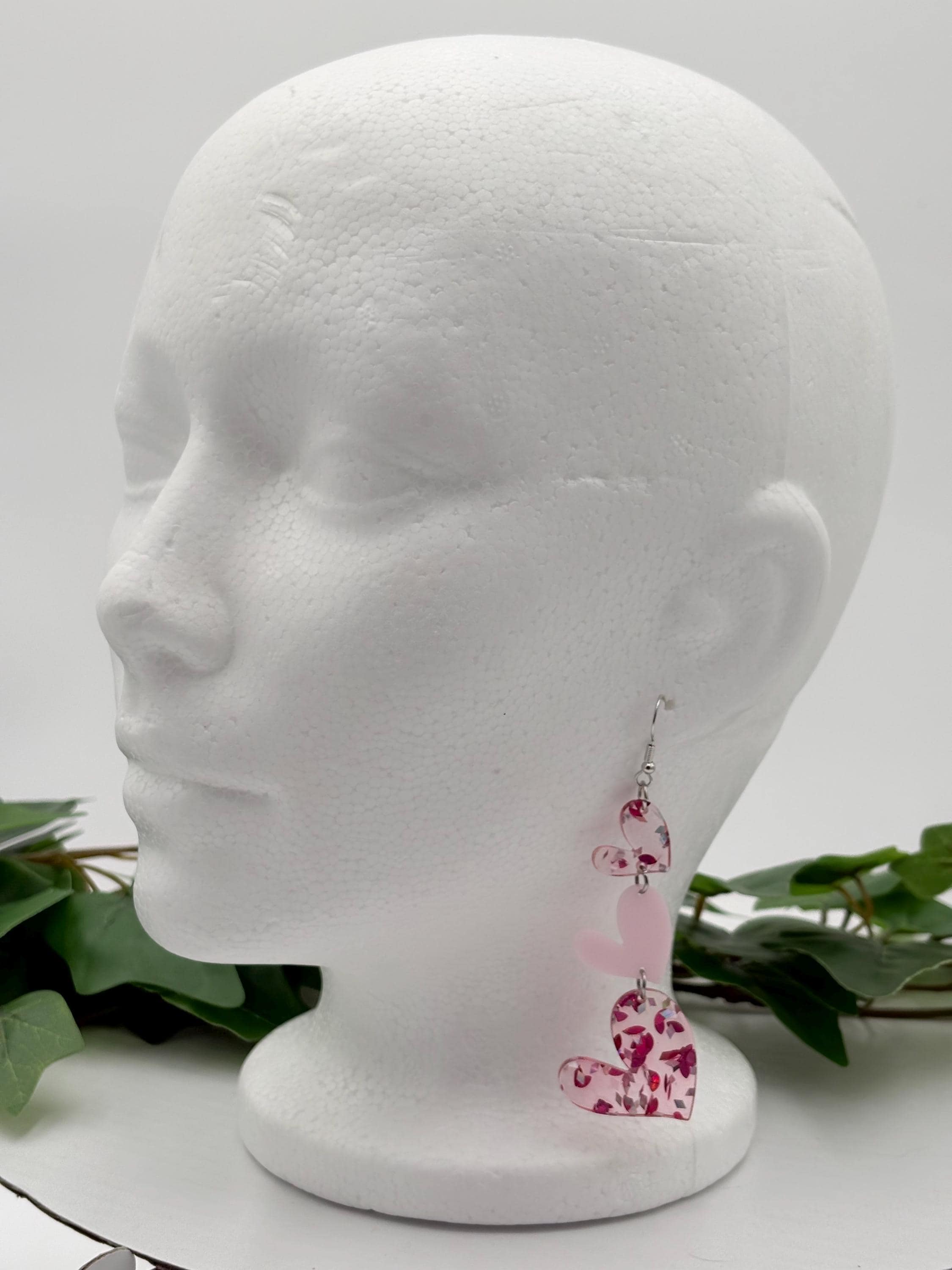 Pink Heart-shaped earrings on a mannequin head with green leaves in the background