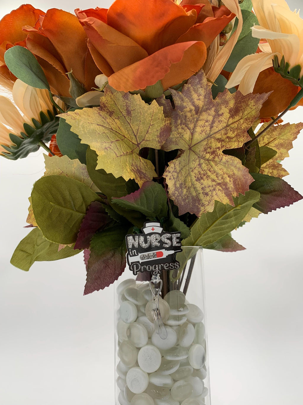 Bouquet of autumn-themed flowers with a 'Nurse in Progress' badge reel in a clear vase on a white background
