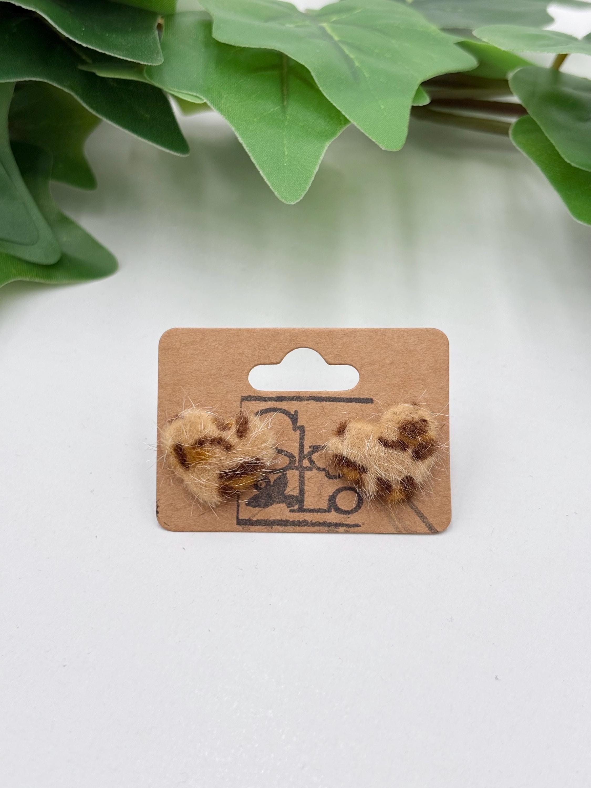 Pair of light brown faux fur leopard print earrings on a cardboard backing with a plant in the background.
