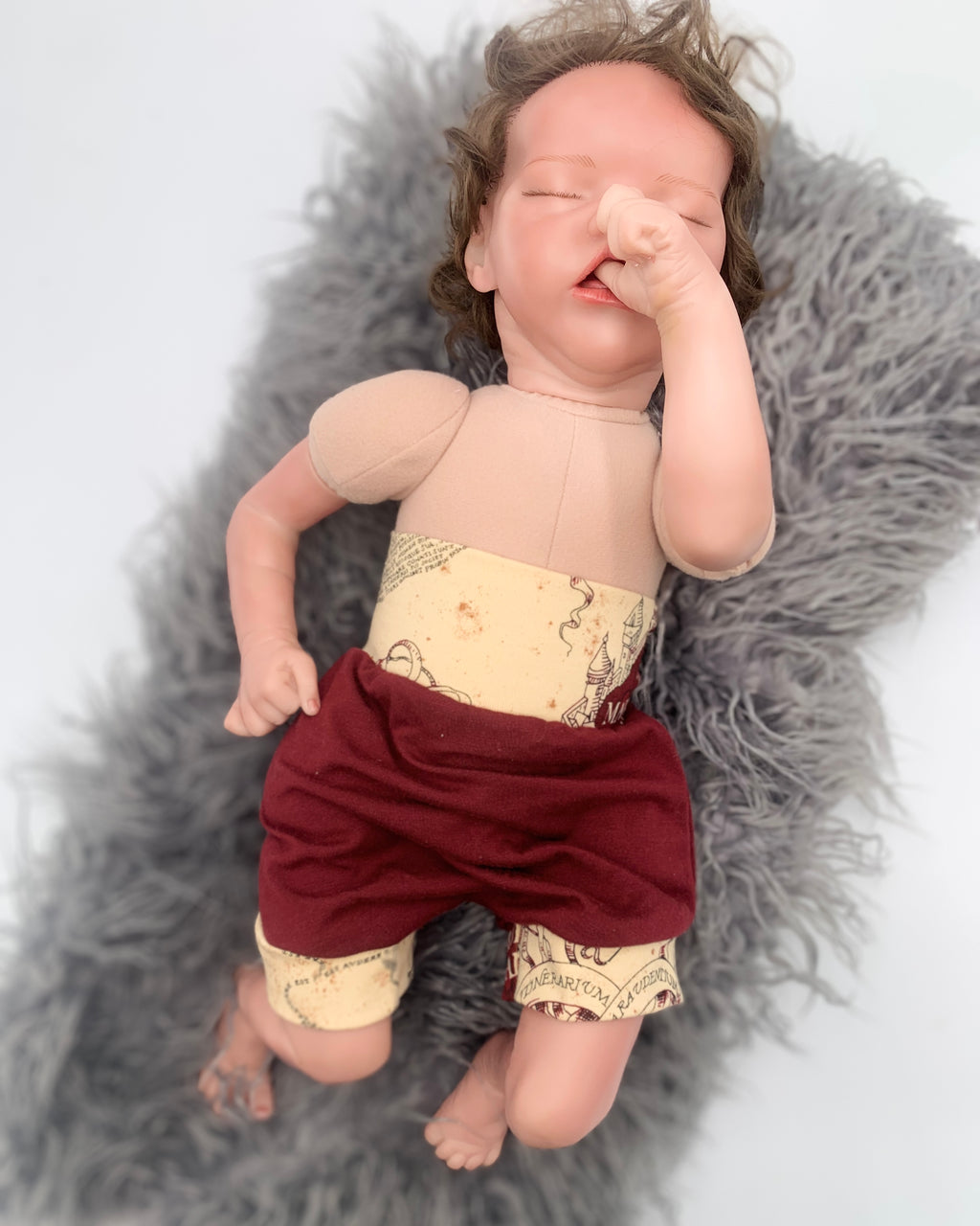 Newborn baby doll wearing Maroon baby bloomers with beige patterned waistband on a white background on a gray fur rug