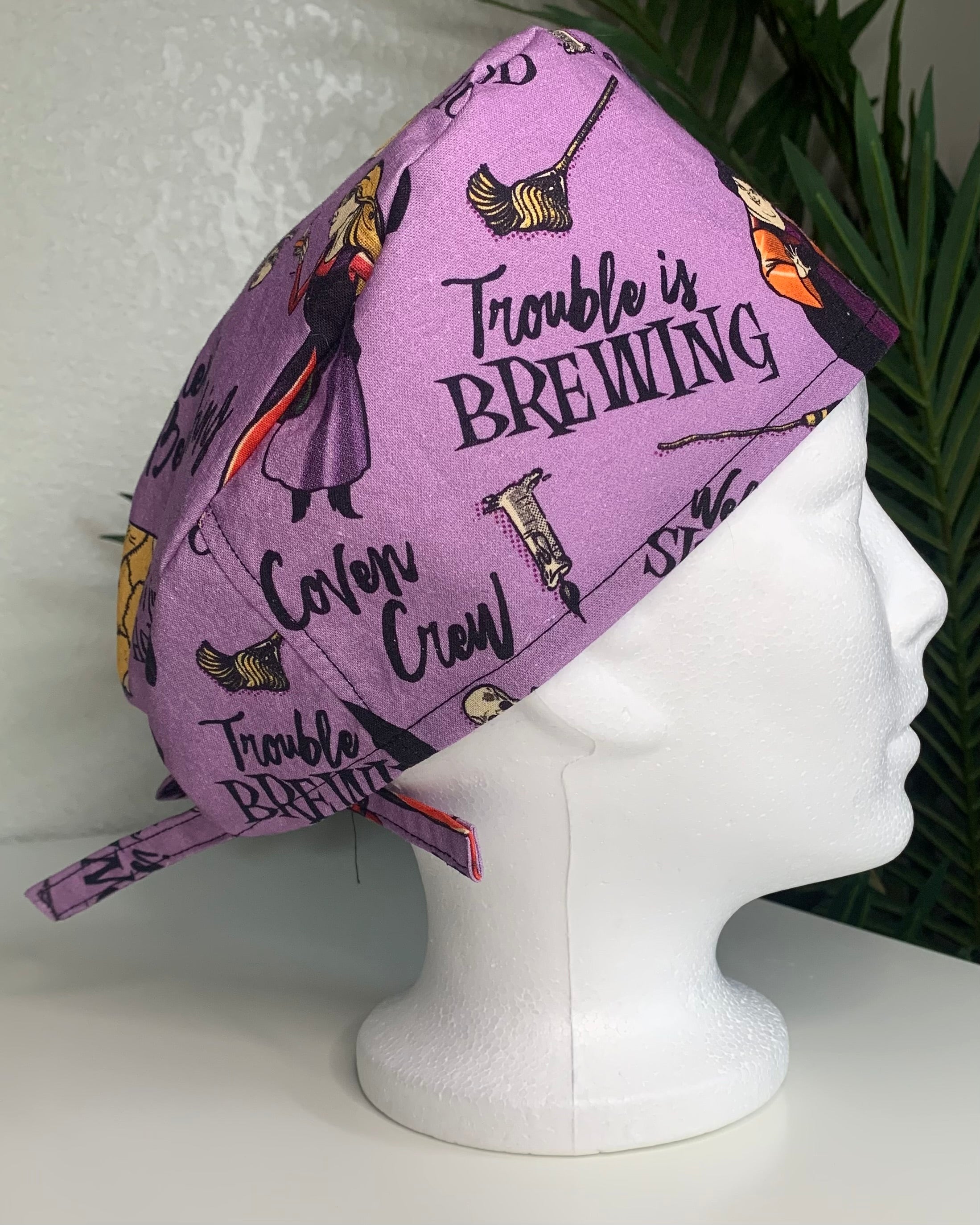 Purple Hocus Pocus patterned scrub cap on a mannequin head with a white background with plant
