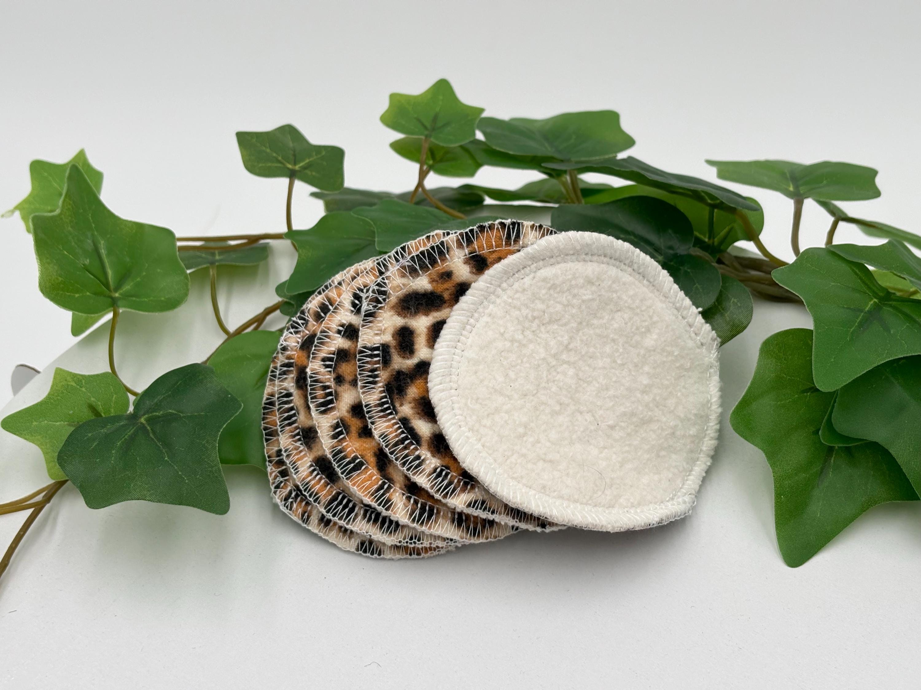 Set of five reusable makeup remover pads with leopard print and white fabric, surrounded by green leaves on a white background.