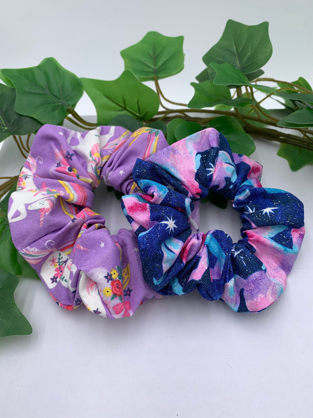Two colorful scrunchies on a white surface with green leaves in the background