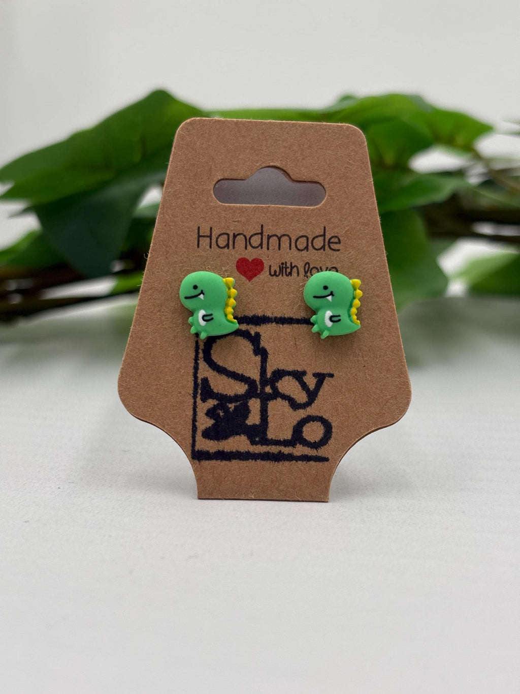 Green dinosaur-shaped earrings on a 'Handmade with Love' card with a blurred green leaf background.