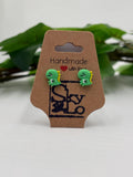 Green dinosaur-shaped earrings on a 'Handmade with Love' card with a blurred green leaf background.