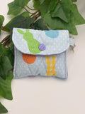 Small quilted headphone holder pouch keychain with colorful easter design on a white surface with green leaves.