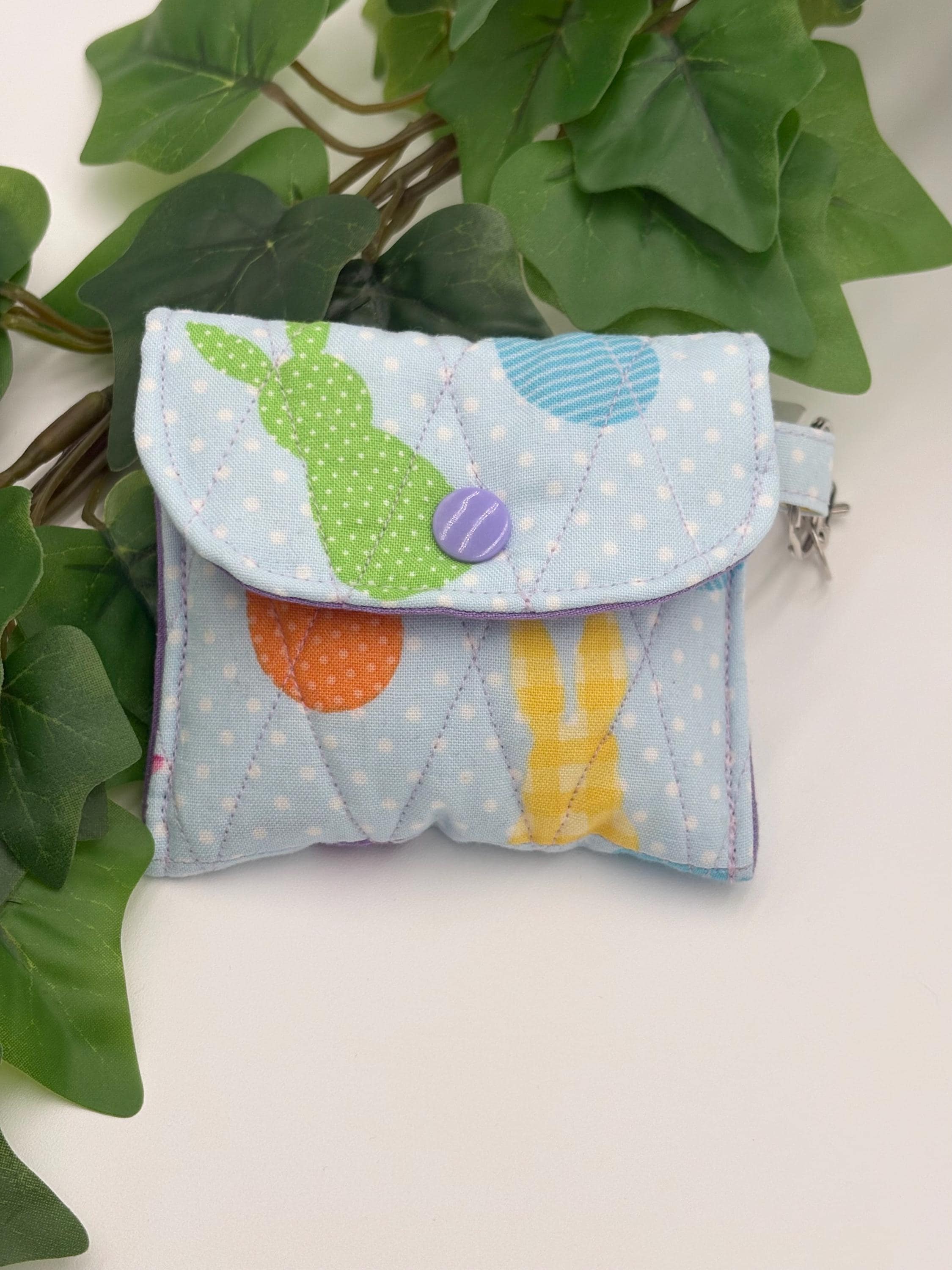 Small quilted headphone holder pouch keychain with colorful easter design on a white surface with green leaves.
