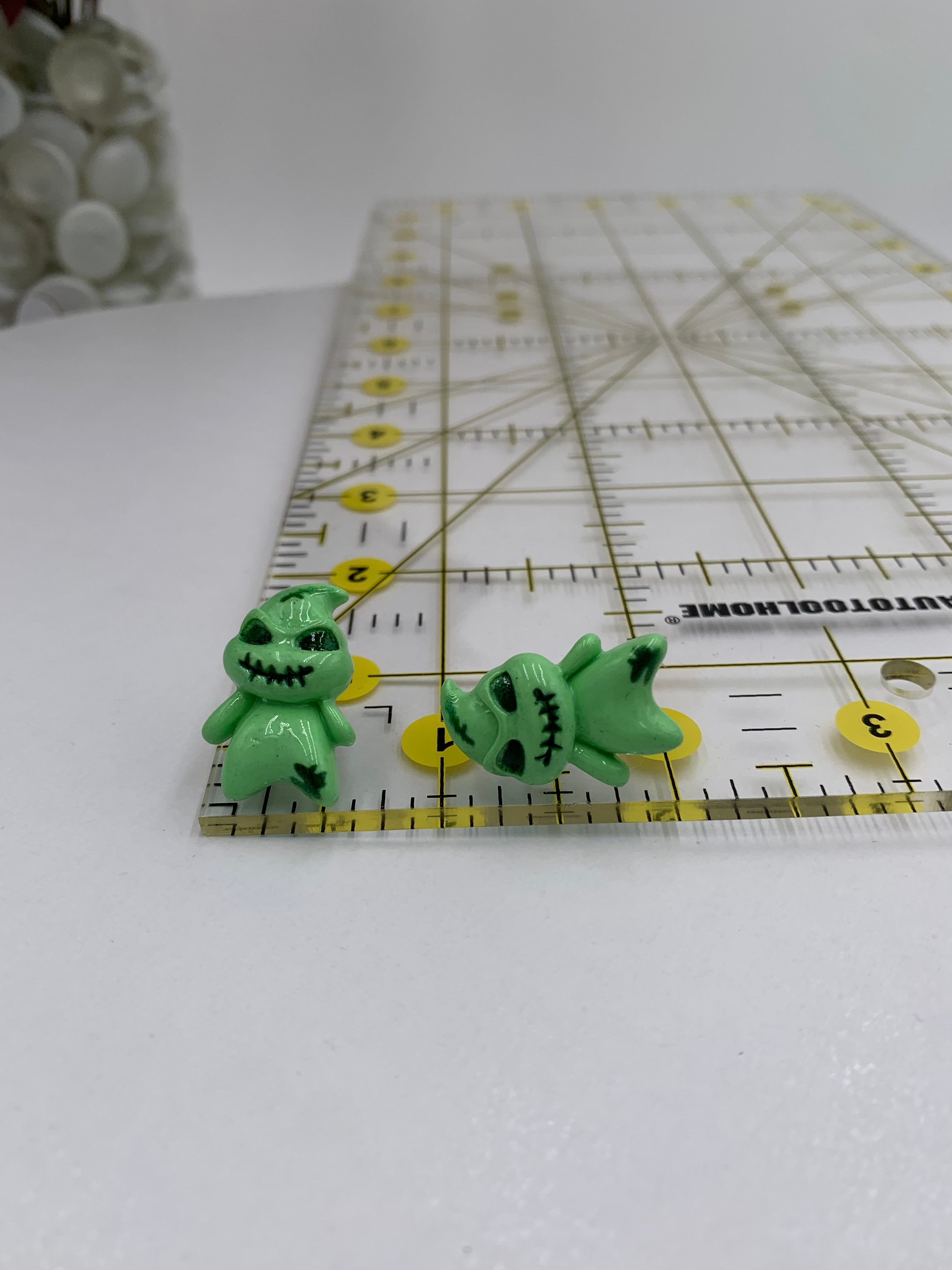 Two green boogeyman-shaped earrings on a ruler against a white background