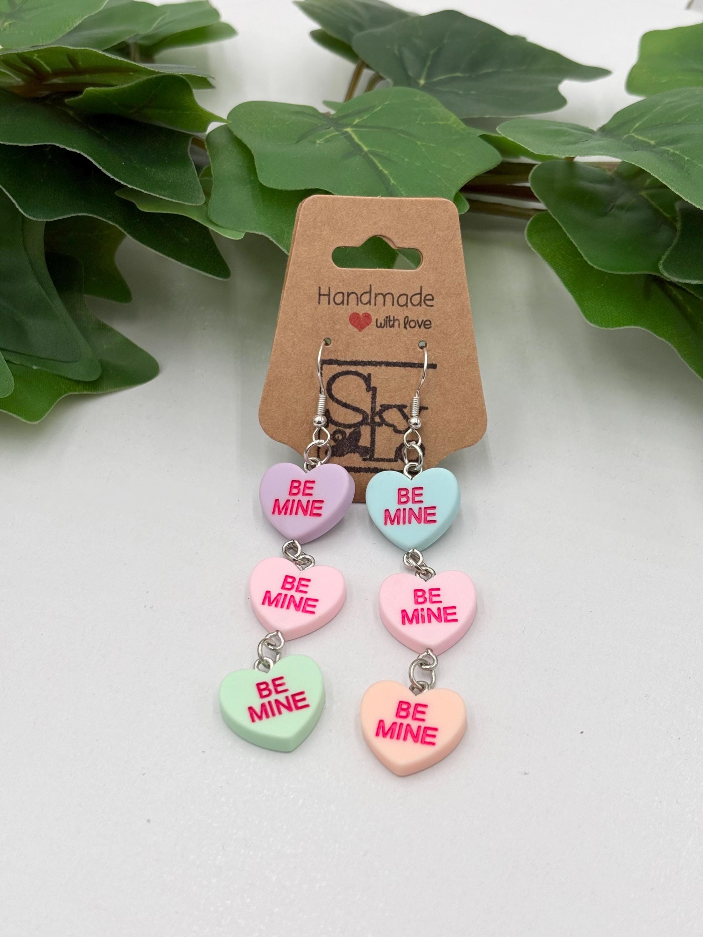 Heart-shaped dangle earrings with 'Be Mine' text on a cardboard backing with green leaves in the background