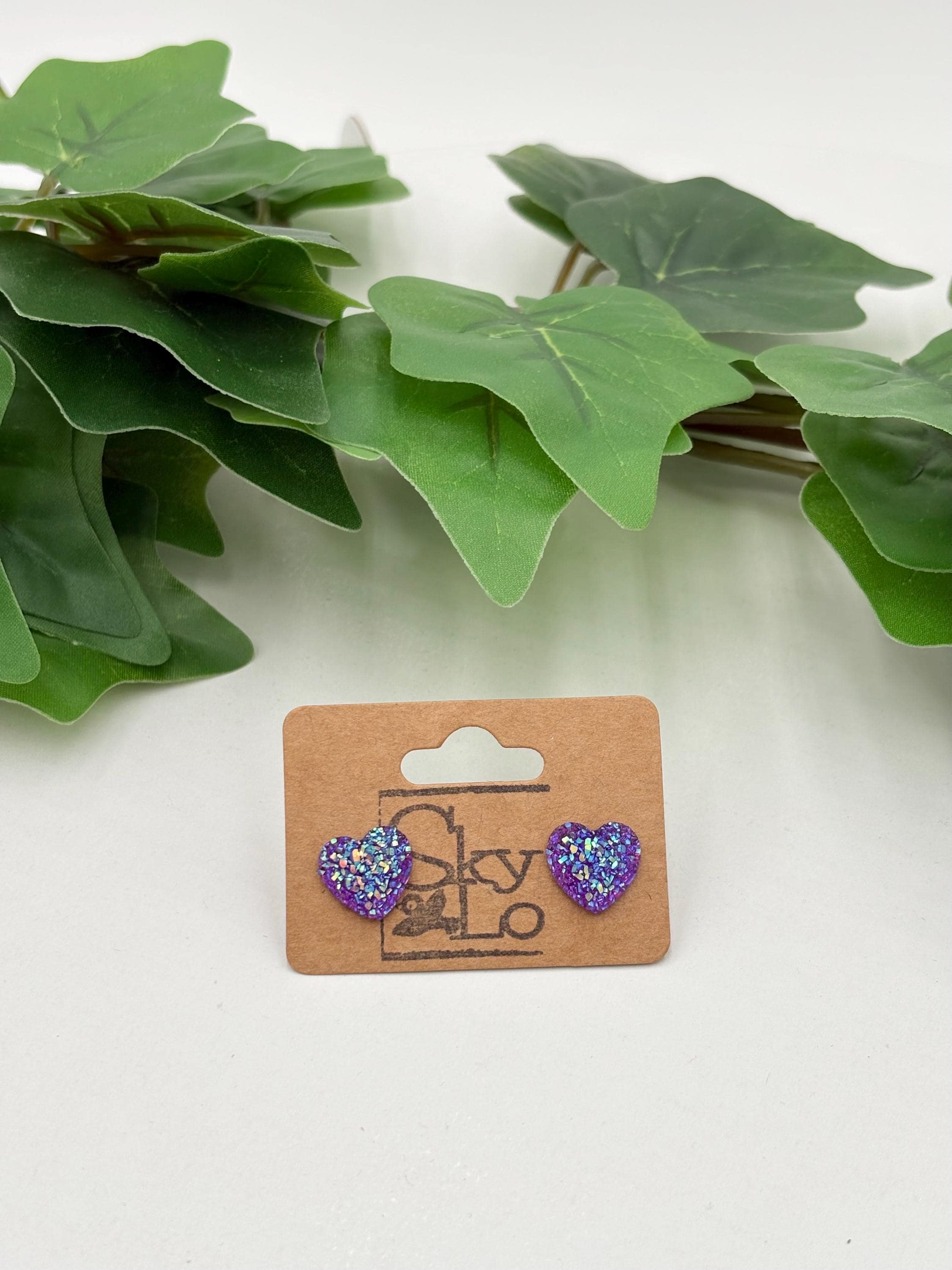 Purple sparkling heart-shaped earrings on a 'SkyLo' branded card with green leaves in the background