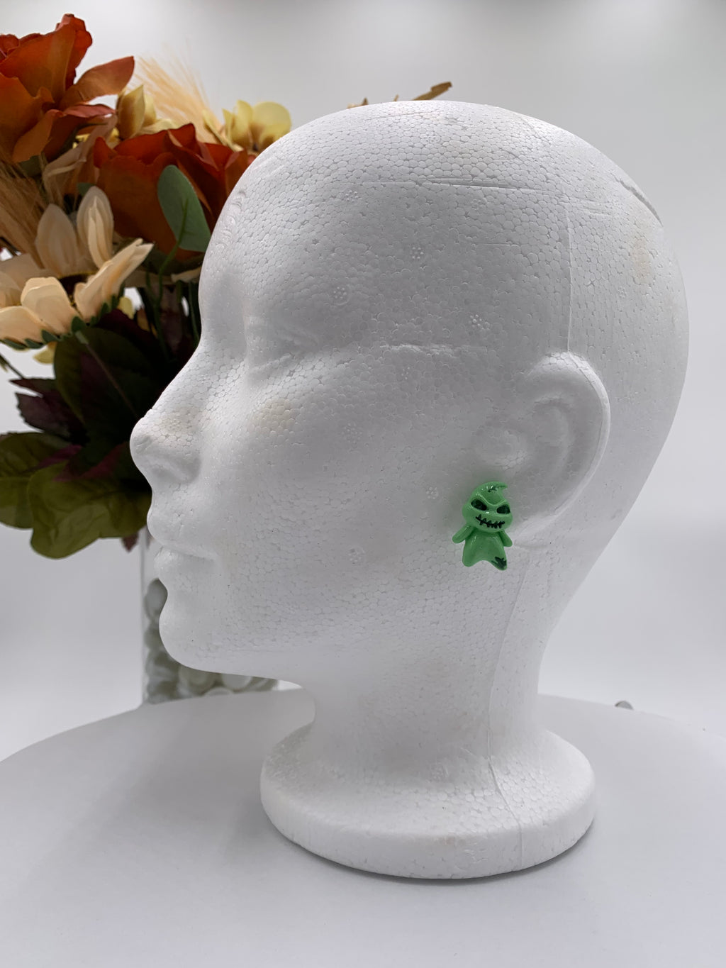 Green boogeyman shaped earring on a white mannequin head with flowers in the background