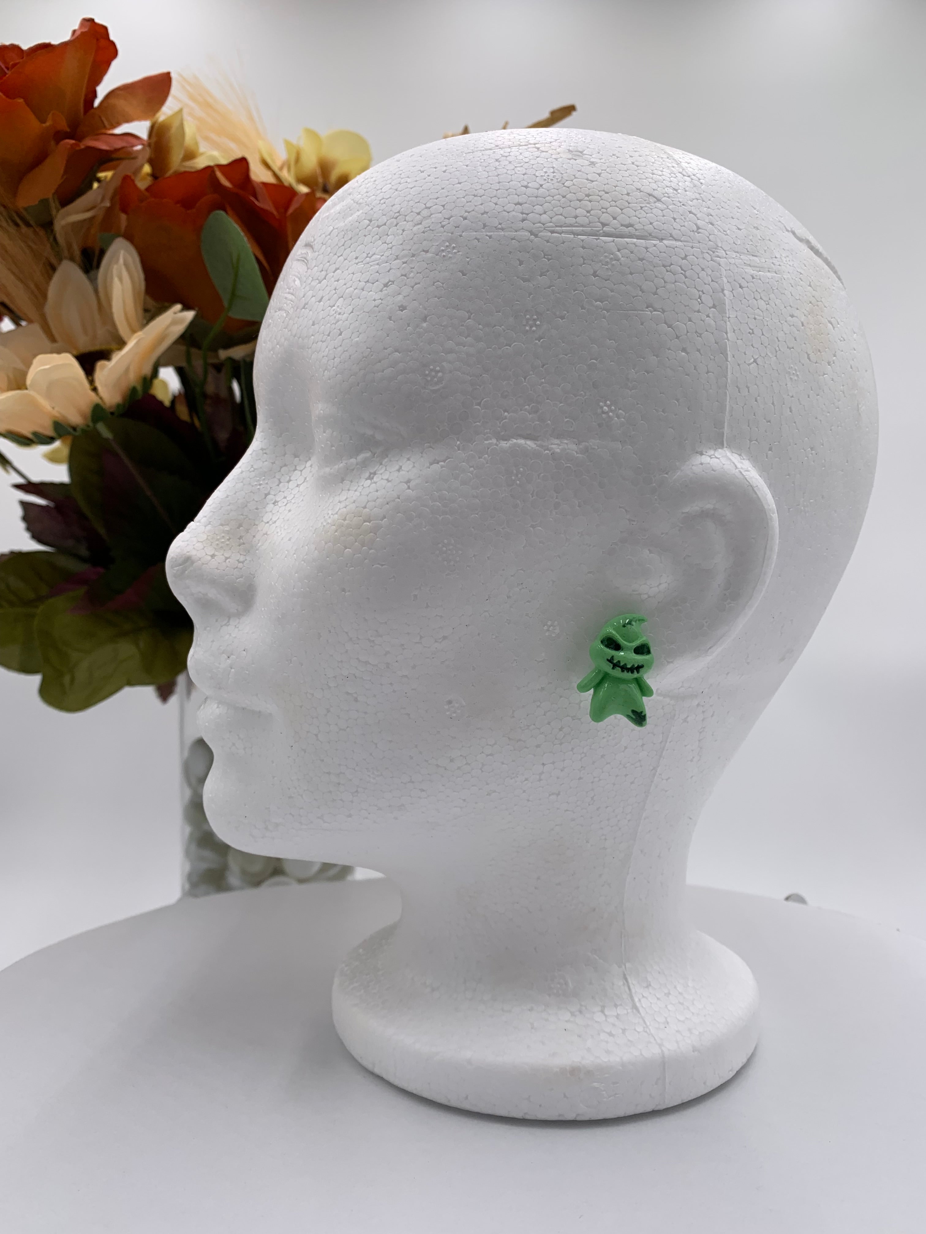 Green boogeyman shaped earring on a white mannequin head with flowers in the background