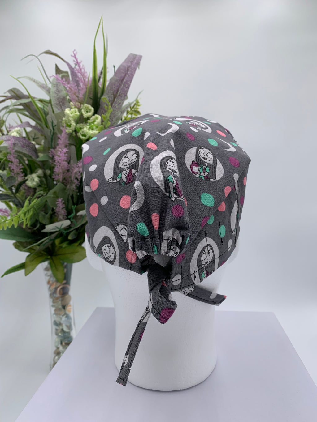 Gray polka dot nightmare before Christmas scrub cap on a mannequin head with flowers, against a white background
