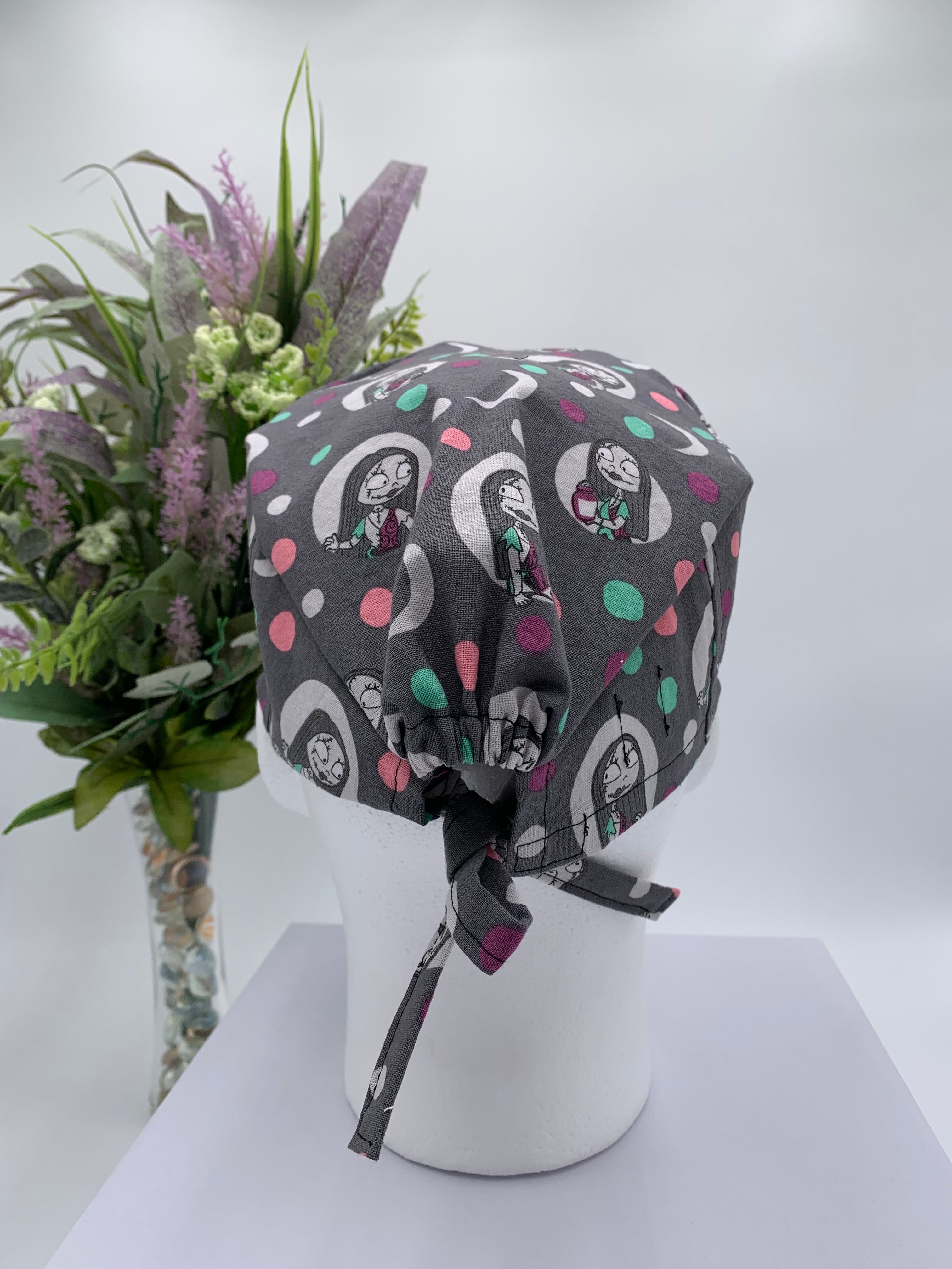 Gray polka dot nightmare before Christmas scrub cap on a mannequin head with flowers, against a white background