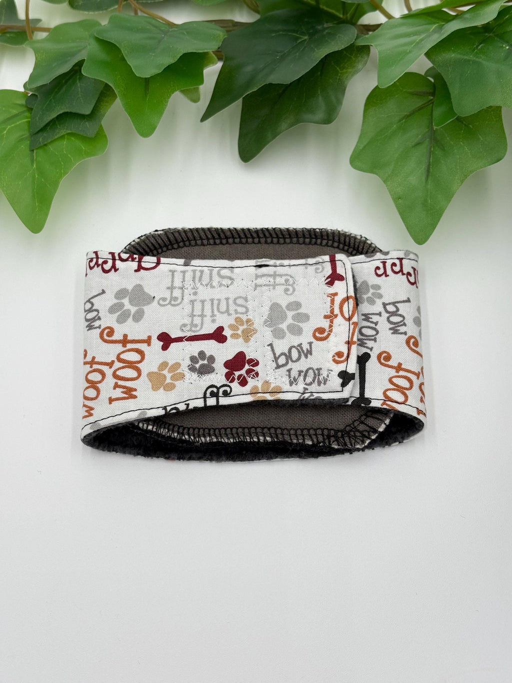 A closed dog belly band with paw prints and 'woof' text with the incontinence liner inside  on a white background with green leaves.