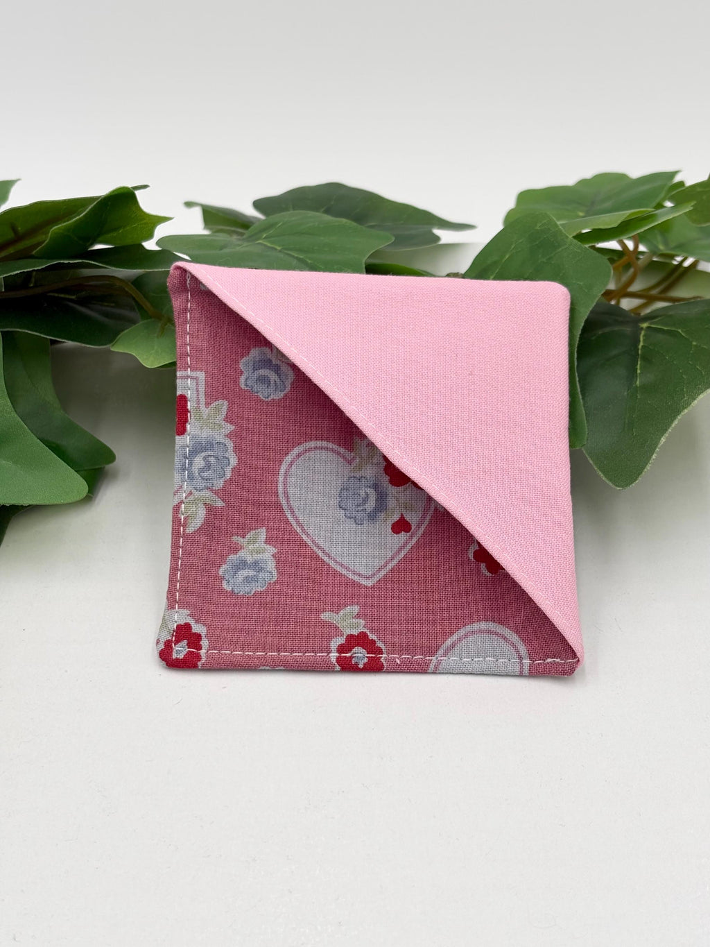 Pink fabric square corner bookmark with hearts and flowers pattern on a white surface with green leaves.