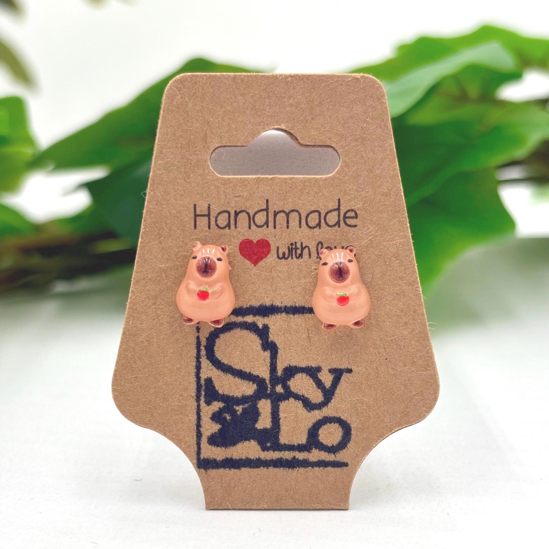 Handmade capybara with red fruit earrings on a branded card with a blurred green plant background