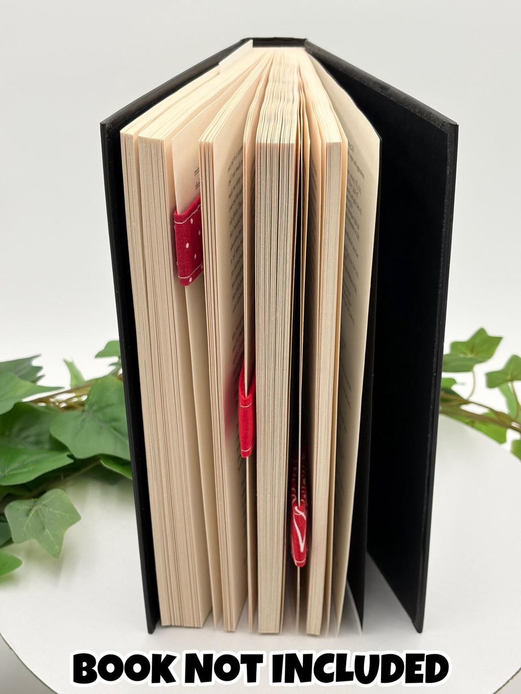 A Book with a green leafy branch on a white surface, text 'BOOK NOT INCLUDED' at the top with three red magnetic bookmarks on pages. 