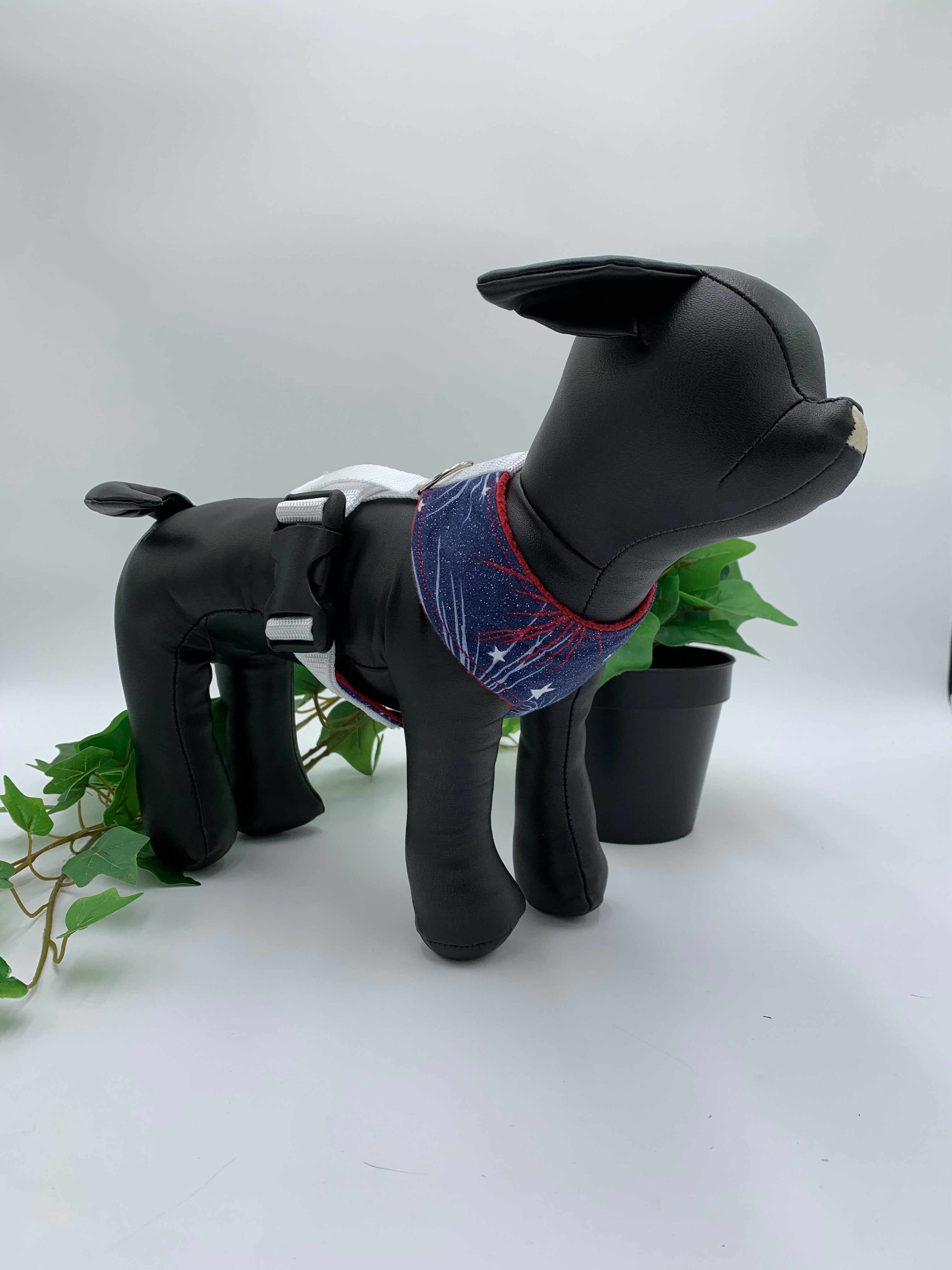 Black dog mannequin with blue 4th of July harness on a white background with a potted green plant