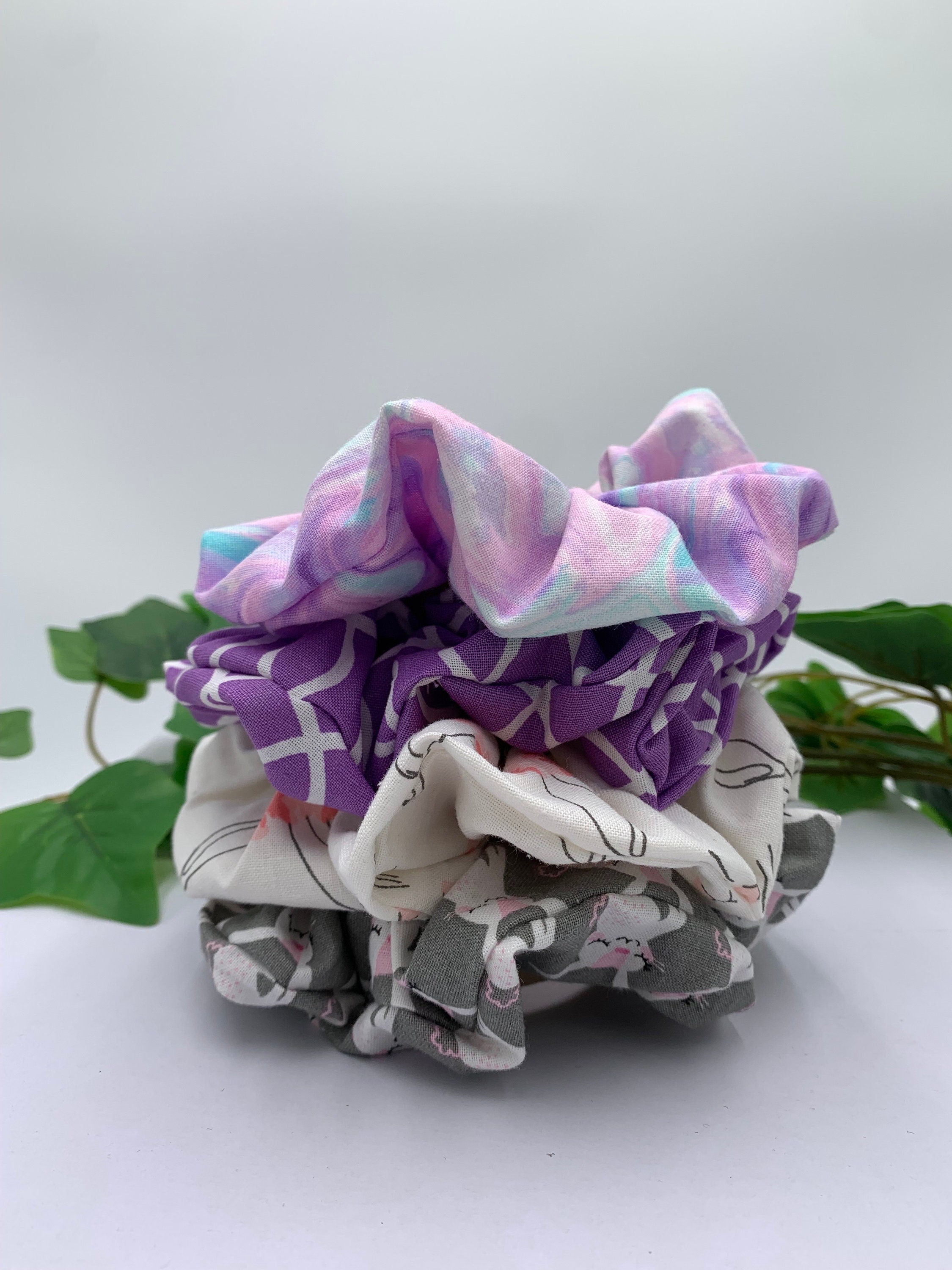 Set of four scrunchies with pastel print, purple and white patterned, white bunny and gray bunny designs on a white background next to green leaves