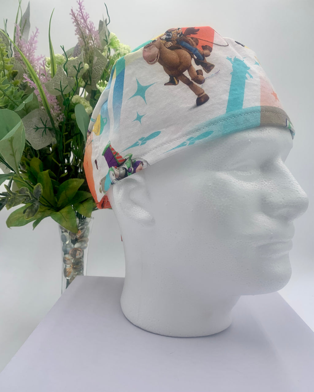 Colorful Toy Story scrub cap on a mannequin head with flowers, against a white background