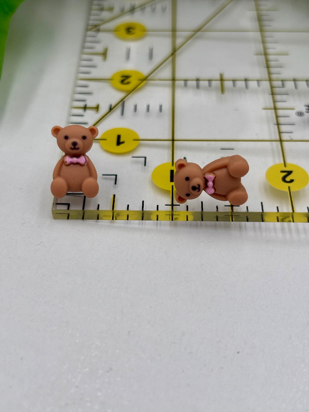 Two small teddy bear earrings with pink bows on a ruler for scale.