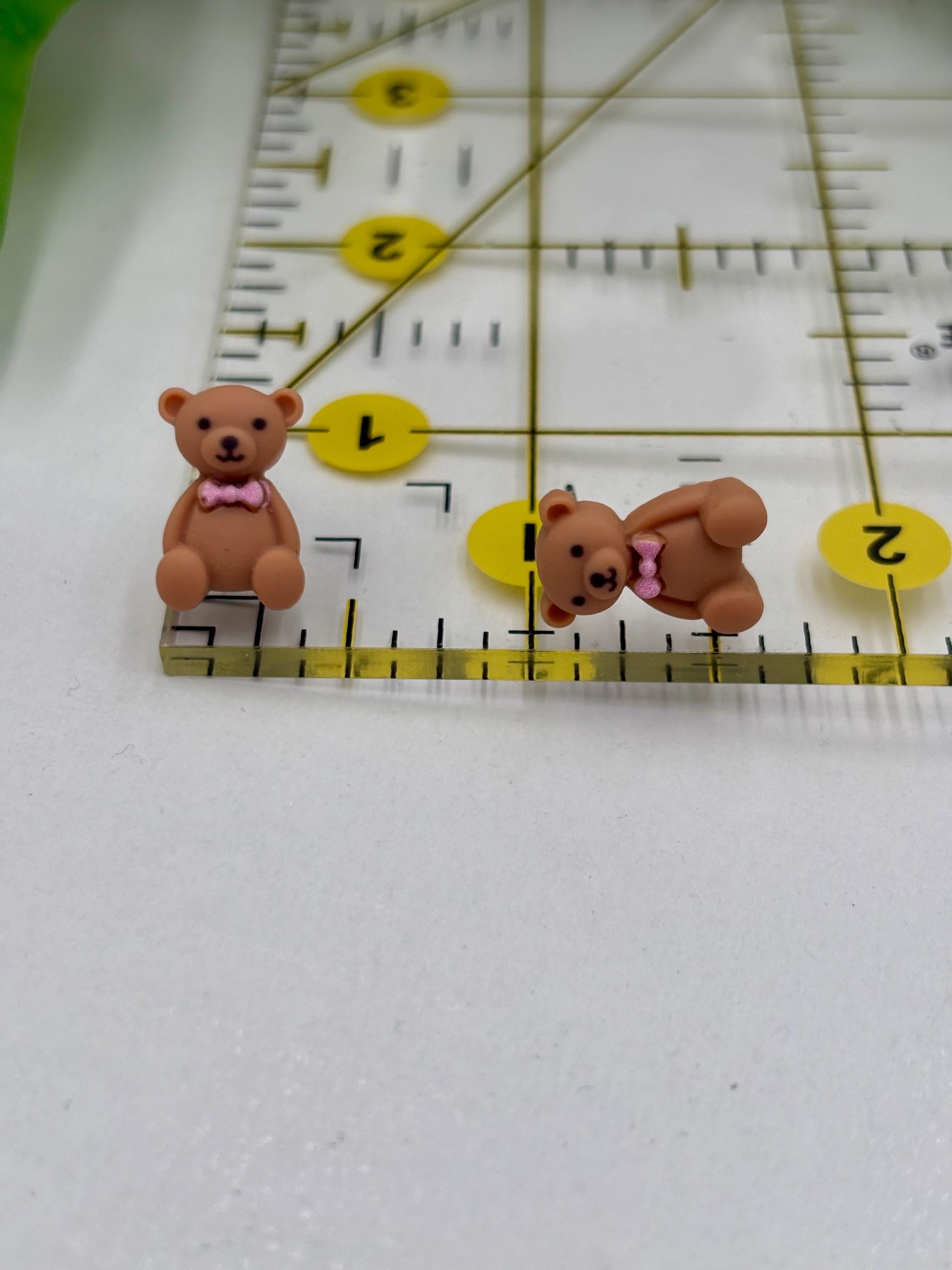 Two small teddy bear earrings with pink bows on a ruler for scale.