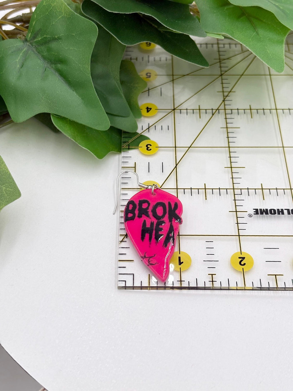 Pink heart shaped earring with 'BROK HEA' text on a white surface with green leaves and a ruler.