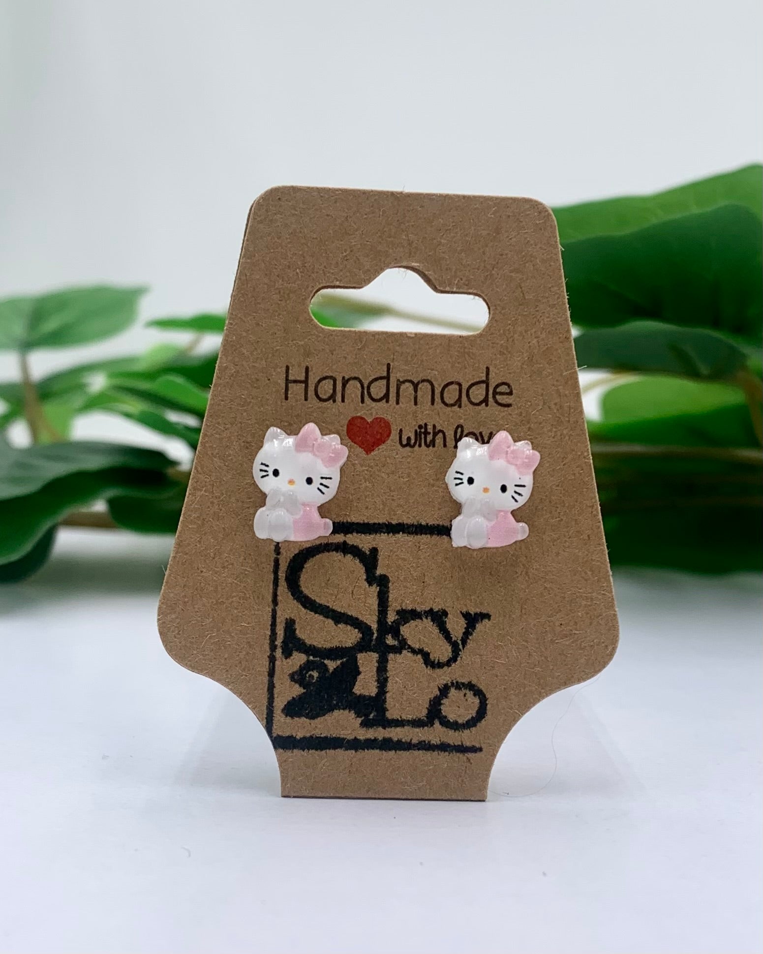 Cat-shaped earrings on a brown card with 'Handmade with Love' text, against a white background with green leaves.