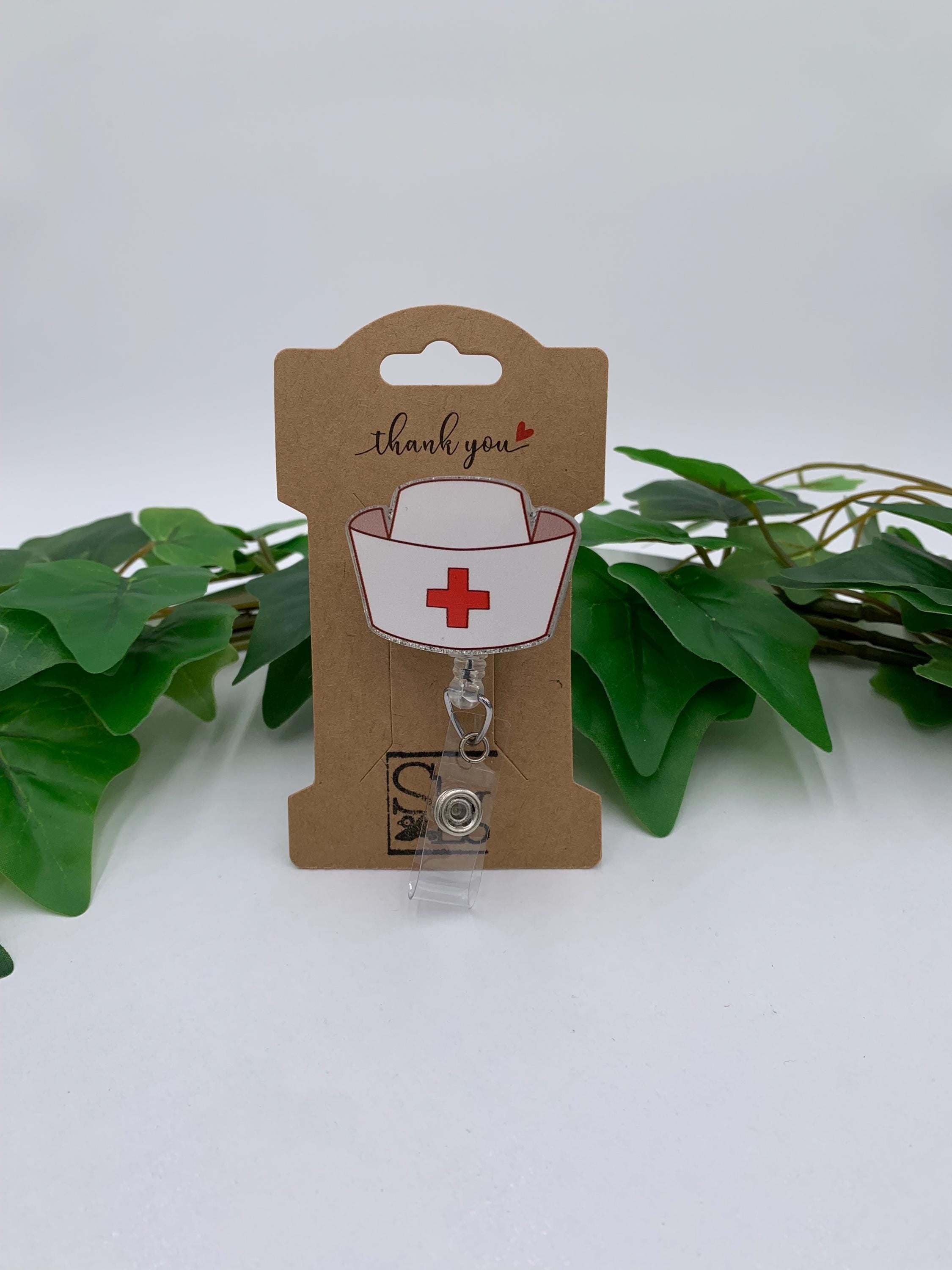 Retractable badge holder with nurse cap design on a cardboard card, surrounded by green leaves.