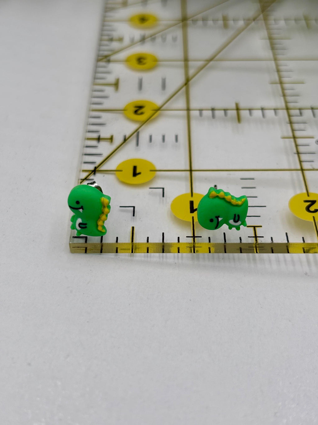 Two green dinosaur earrings next to a ruler on a white background