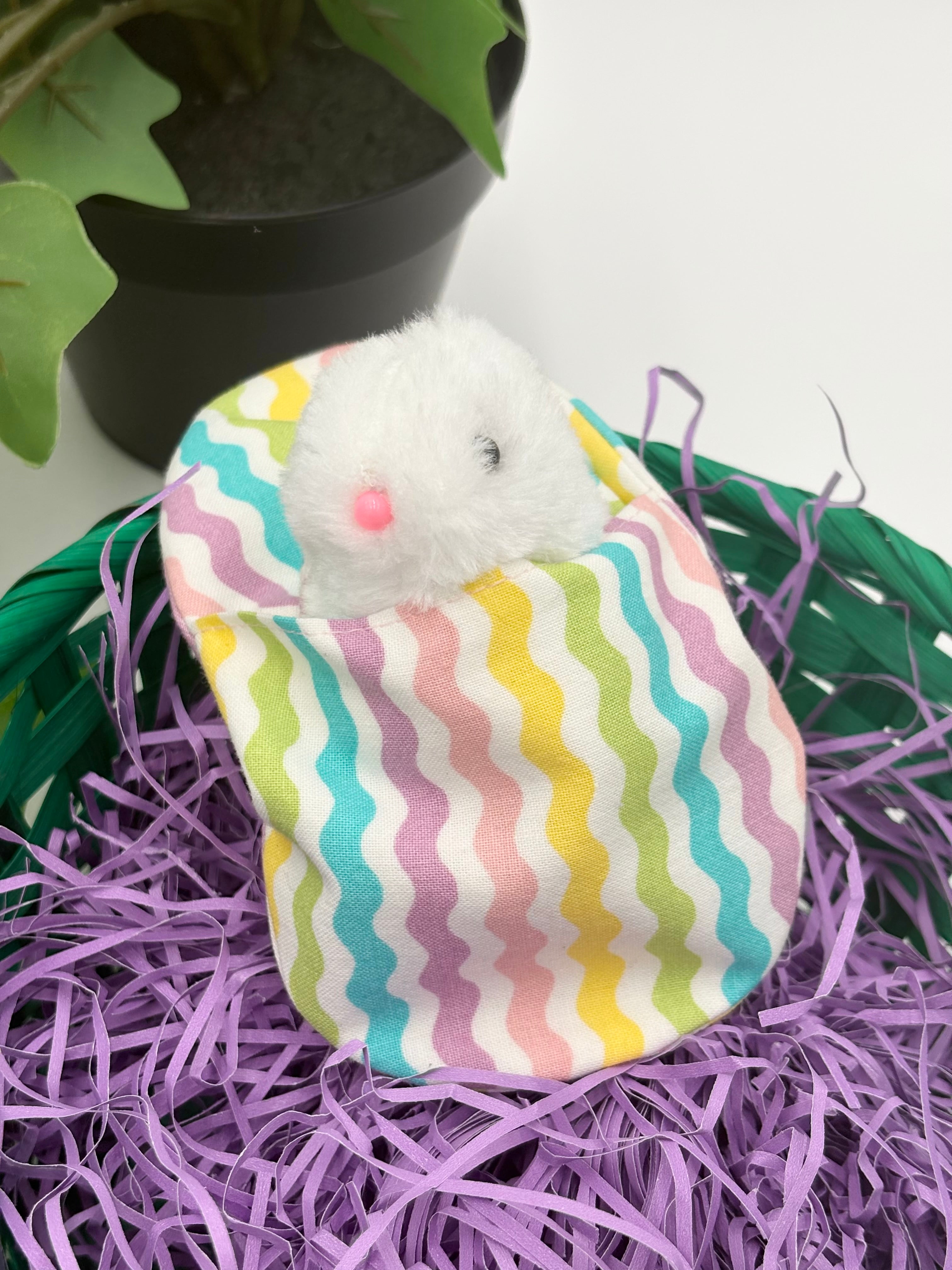 Small plush toy bunny in a colorful striped Easter egg filler on purple Easter grass in a green basket with a potted plant in a white background