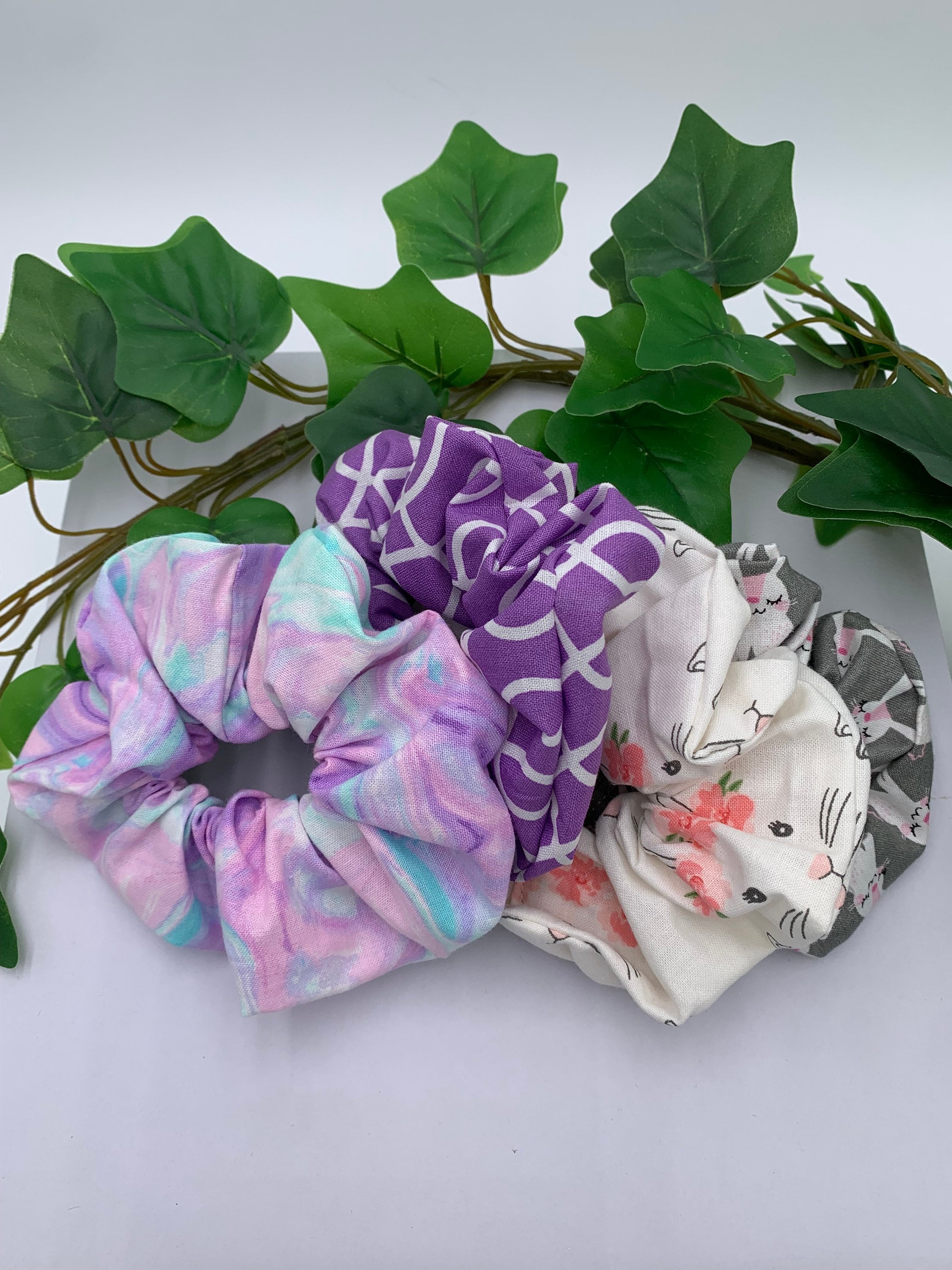 Set of four scrunchies with pastel print, purple and white patterned, white bunny and gray bunny designs on a white background next to green leaves