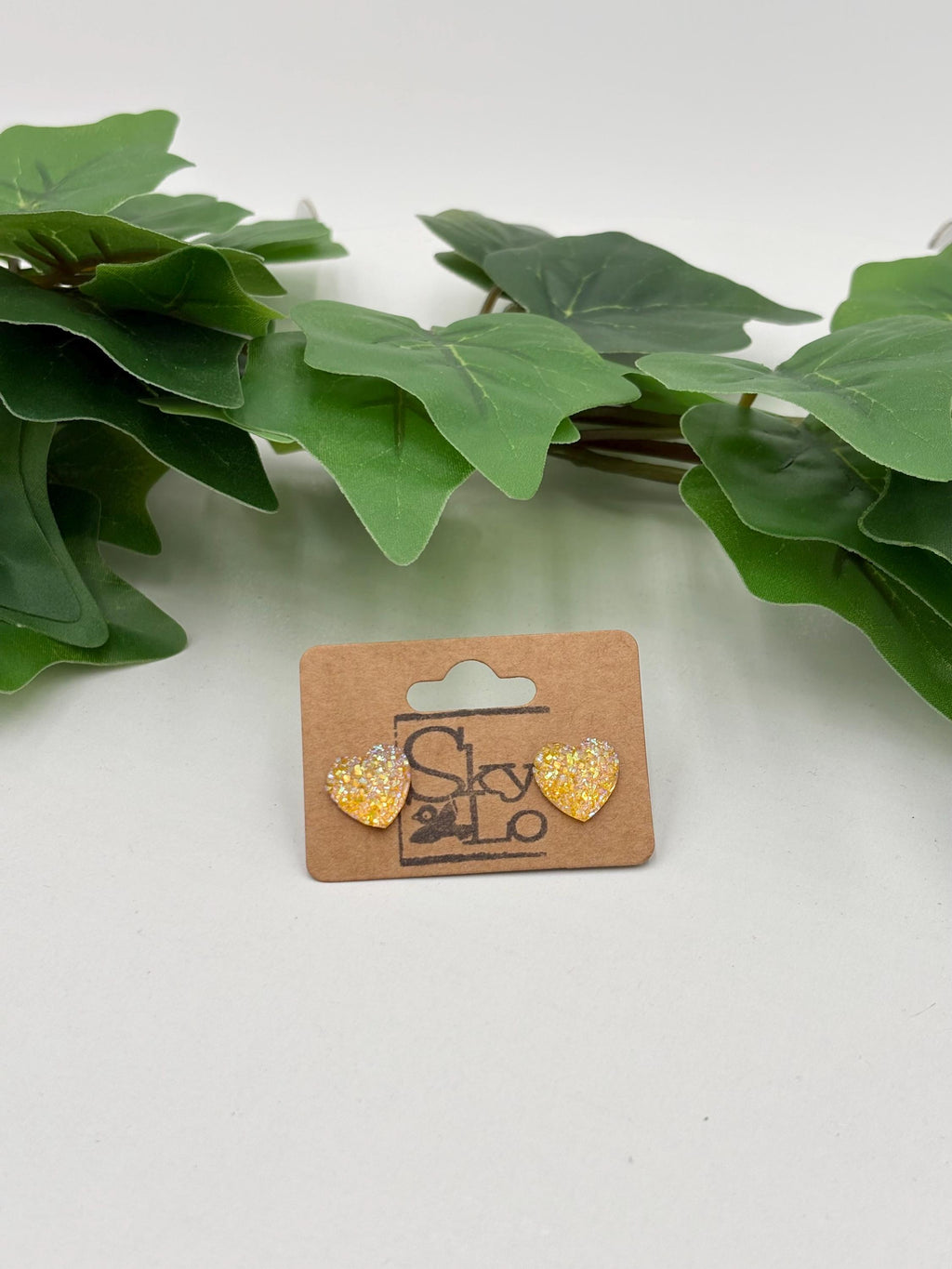 Yellow sparkling heart-shaped earrings on a 'SkyLo' branded card with green leaves in the background